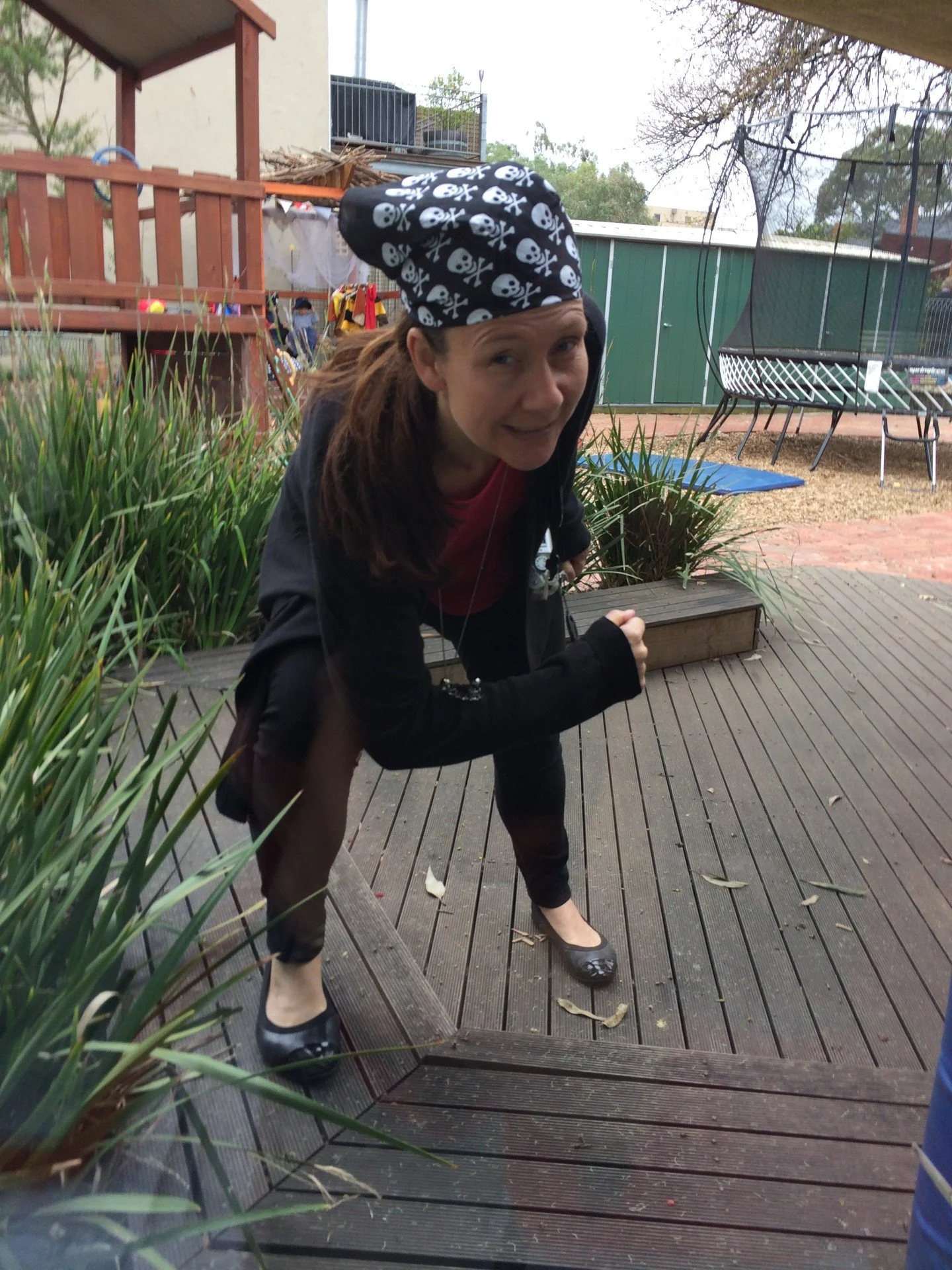 Woman bending forward on a wooden deck, wearing a black jacket, black shoes, and a skull and crossbones bandana, outdoors with plants and outdoor furniture visible.