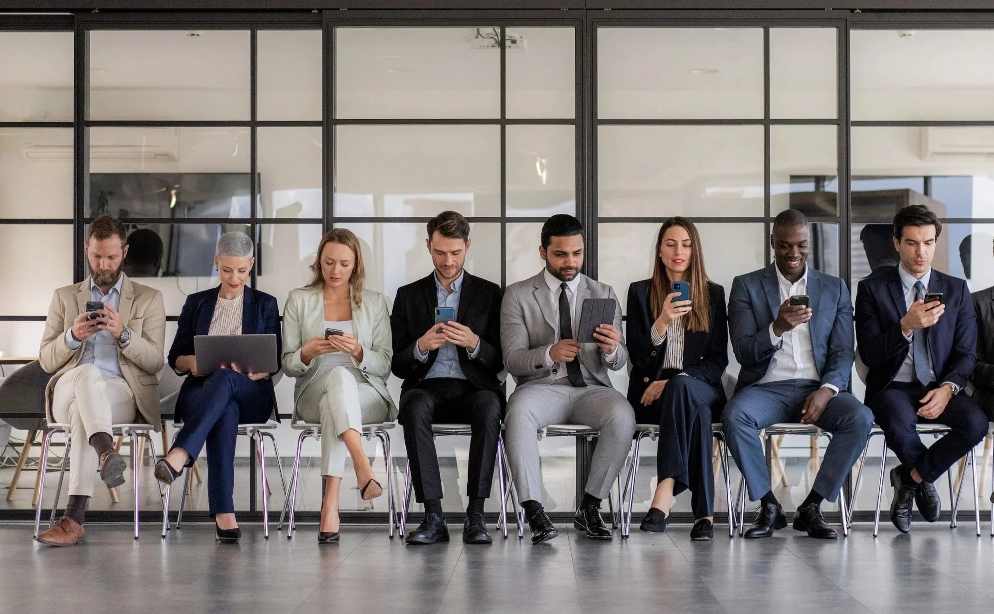 Eight business professionals sitting on chairs in a modern office lobby, all engaged with their smartphones or devices.