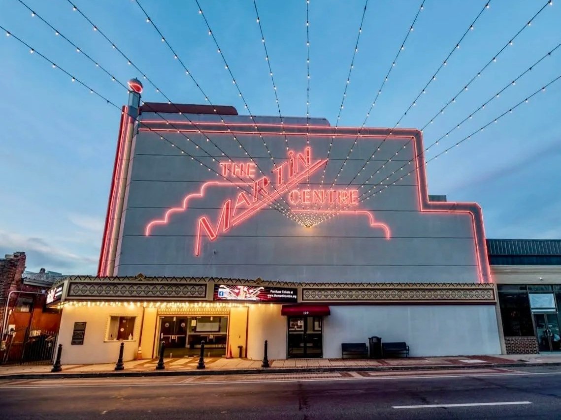 Front view of The Majestic Theatre building with pink neon sign and decorative lights overhead, with a ticket booth and benches outside.