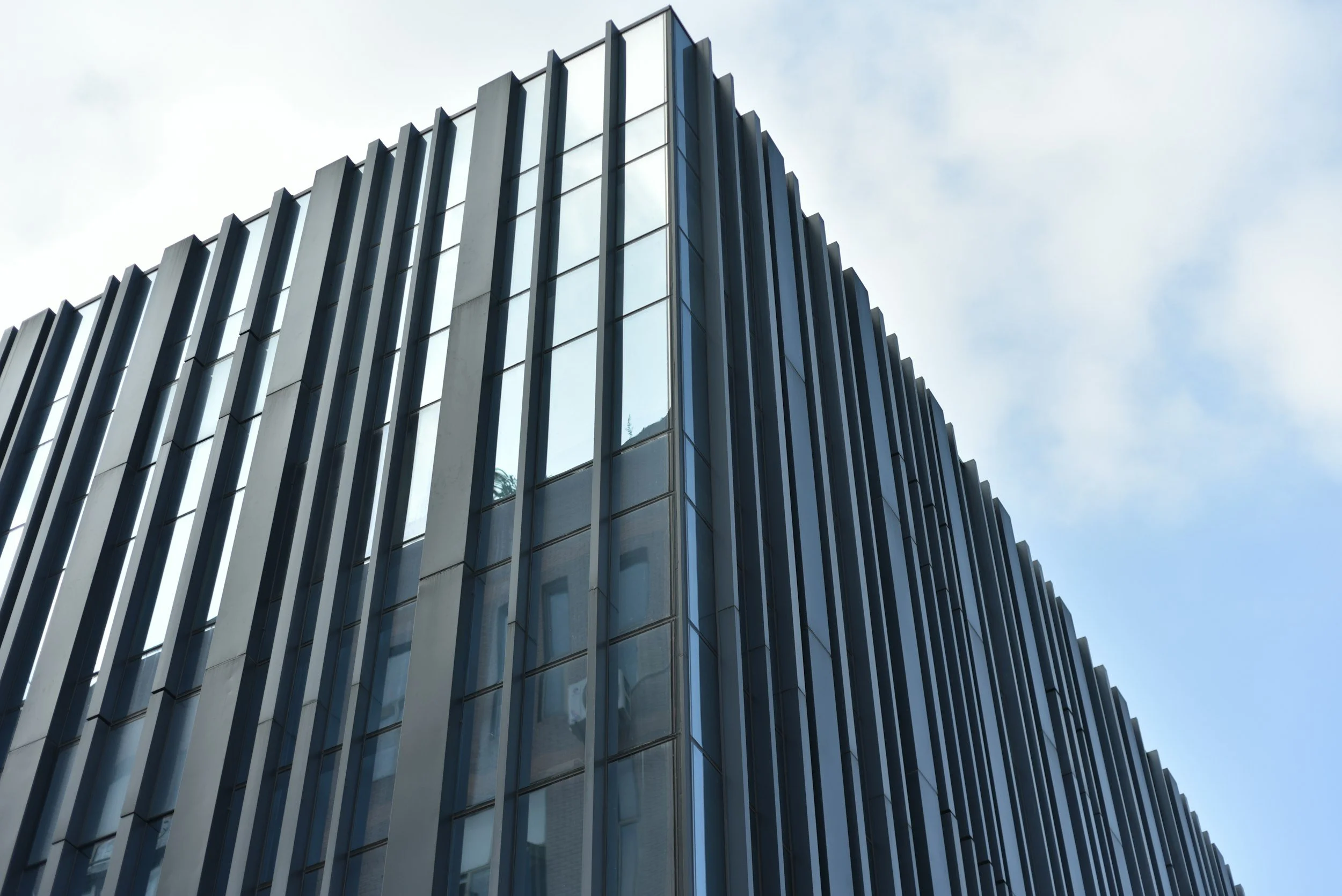 A modern building with a facade made of vertical metal slats and reflective glass windows, set against a blue sky with some clouds.