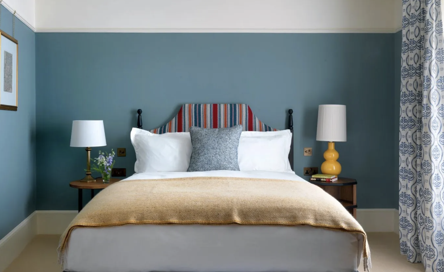 A neatly made bed with white linens, a colorful striped headboard, a gray throw pillow, and a beige blanket. There are two bedside tables with lamps, one white and one yellow, and a window with patterned curtains.