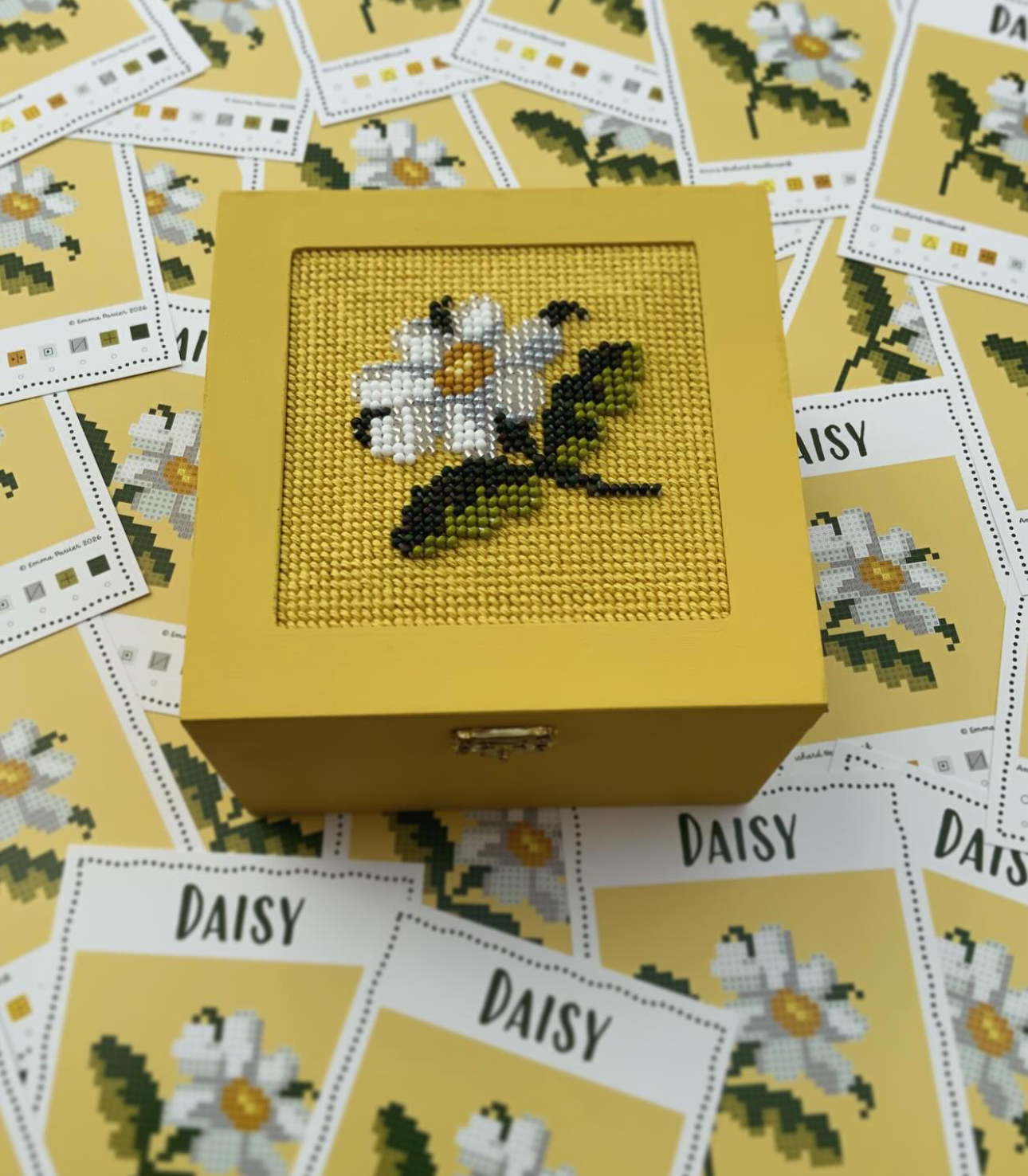 A yellow jewelry box with a cross-stitched gerbera daisy inside, surrounded by Daisy-themed cross-stitch patterns on paper.