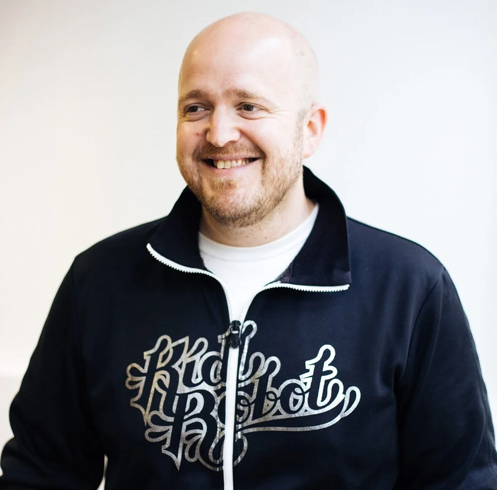 A smiling, bald man with a beard, wearing a black zip-up jacket with white lettering, against a plain light-colored background.