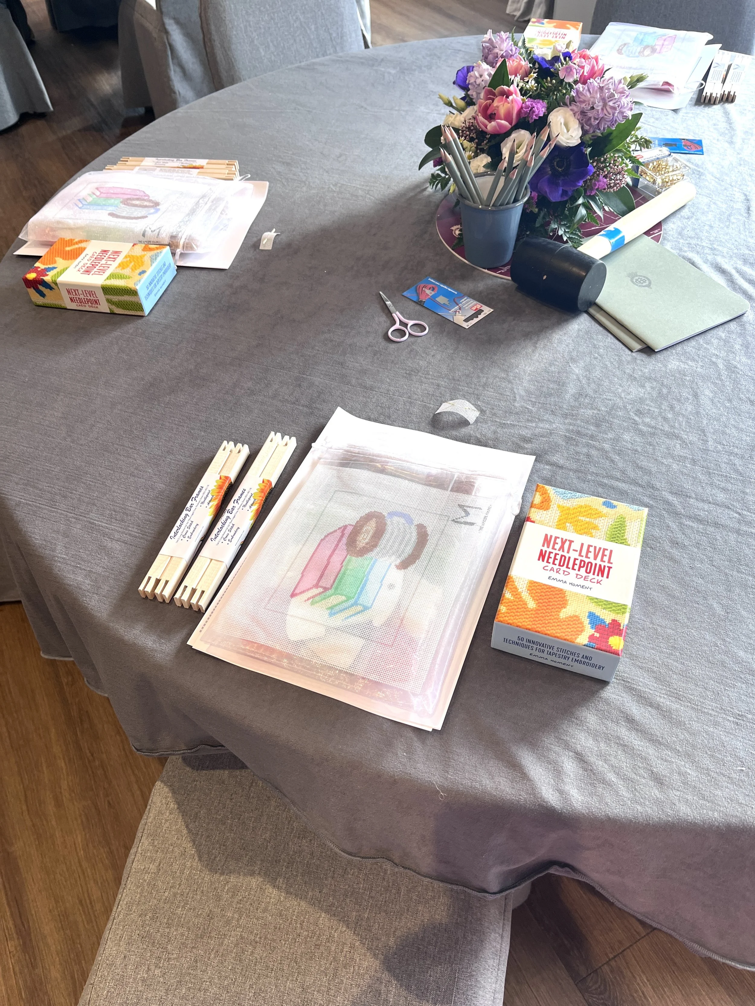 A round table covered with a gray cloth, decorated with a bouquet of pink, purple, and white flowers in a blue vase. The table has several craft supplies including a stack of embroidery cross-stitch kits, a pack of fabric, a pair of scissors, pens, a box of needles, a rolled-up paper, and a stack of napkins. There is also a rectangular transparent embroidery tray with a partially completed cross-stitch pattern. The background shows chairs around the table and a wooden floor.