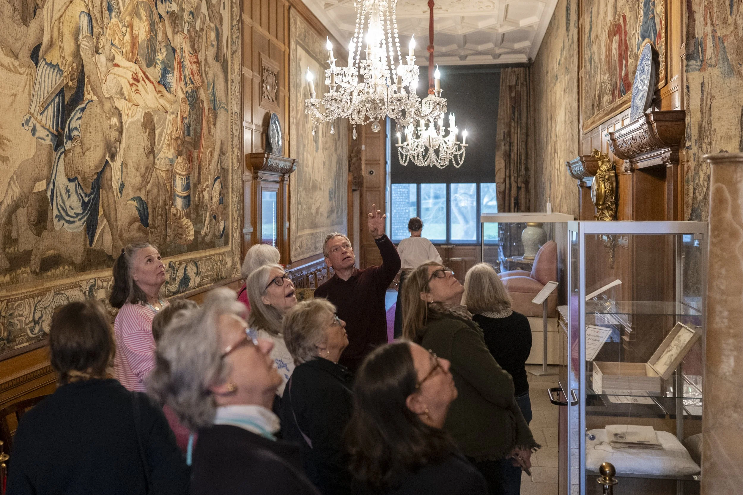 A group of people sit and look up at an ornate chandelier hanging from an elaborately decorated ceiling in a room with wooden walls and large tapestries. Some individuals are pointing or looking towards a display case.