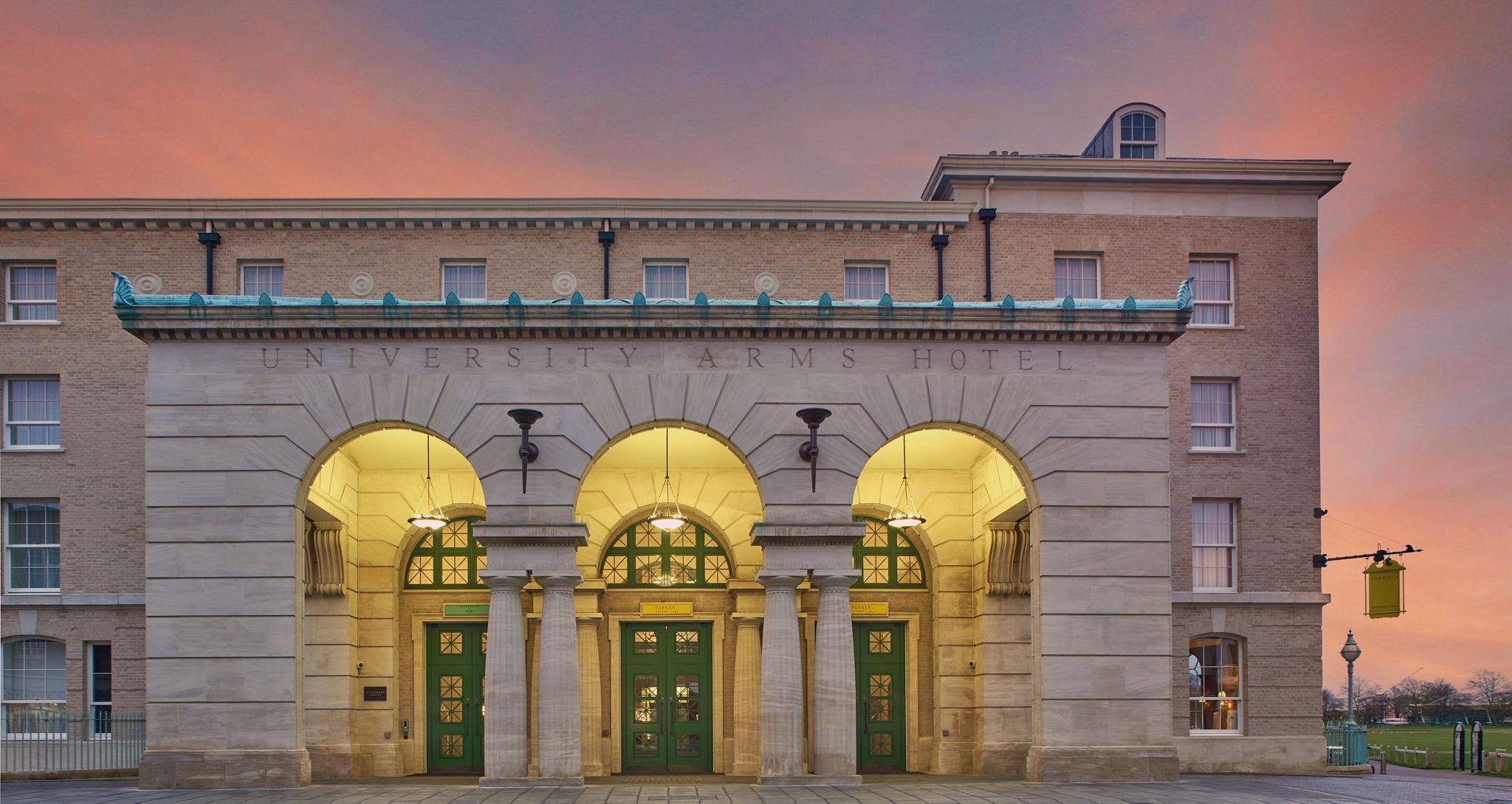 The University Arms Hotel entrance at dusk with glowing yellow lights illuminating the arched portico and green double doors, against a sunset sky.