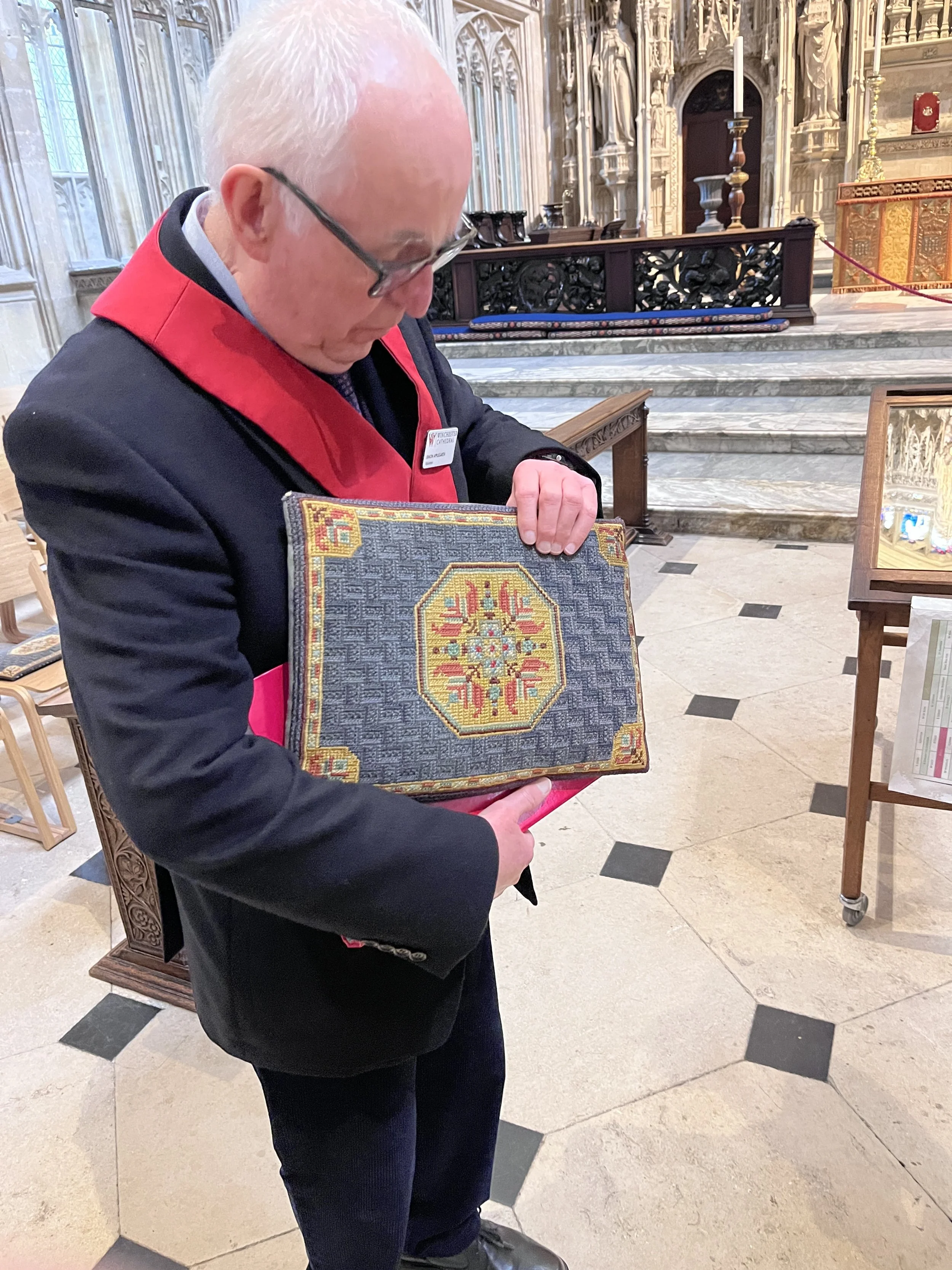 A man with glasses and white hair is standing in a church, holding a framed embroidery piece with a coat of arms, looking down at it.