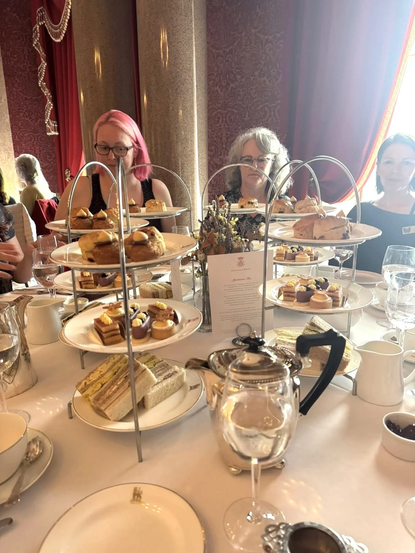 Elegant afternoon tea setting with tiered trays of assorted baked goods, finger sandwiches, cakes, and pastries, glasses of water, teapots, and fine china on a white tablecloth, in a luxurious room with red and gold curtains.