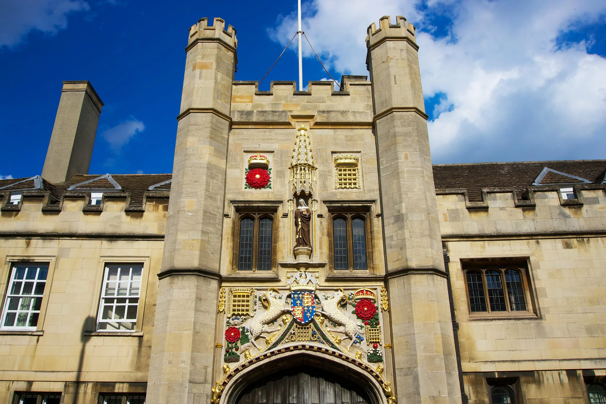 Front view of a historic stone castle-like building with a central tower, decorative coat of arms, statues, and ornate gold embellishments, set against a blue sky with some clouds.