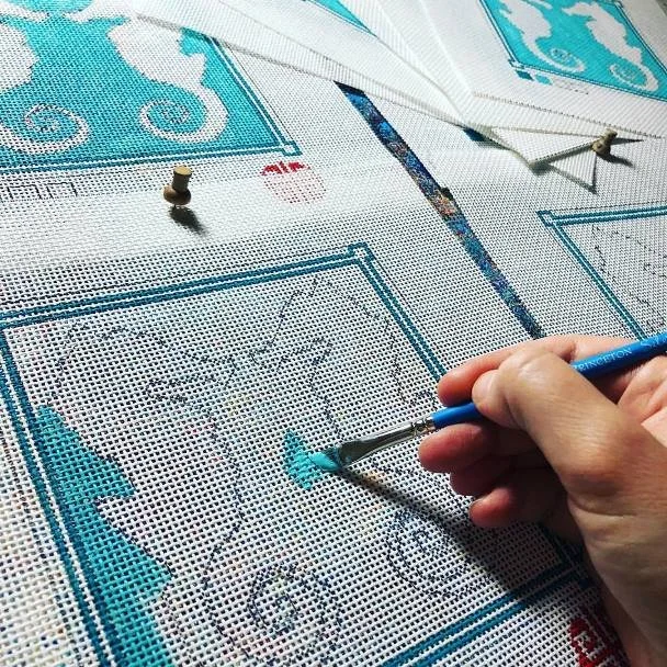 Close-up of a person cross-stitching a blue and white floral pattern on Aida cloth with embroidery tools and finished sections visible.