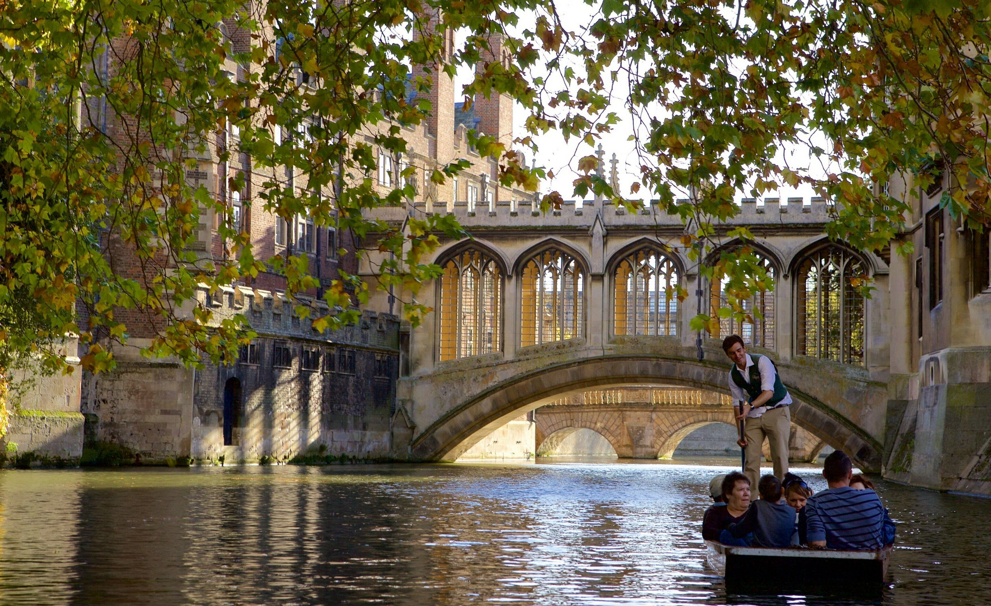 A group of people in a boat on a river beneath a historic stone bridge, with an ornate building with tall windows and a tower partially obscured by green tree leaves in the background.