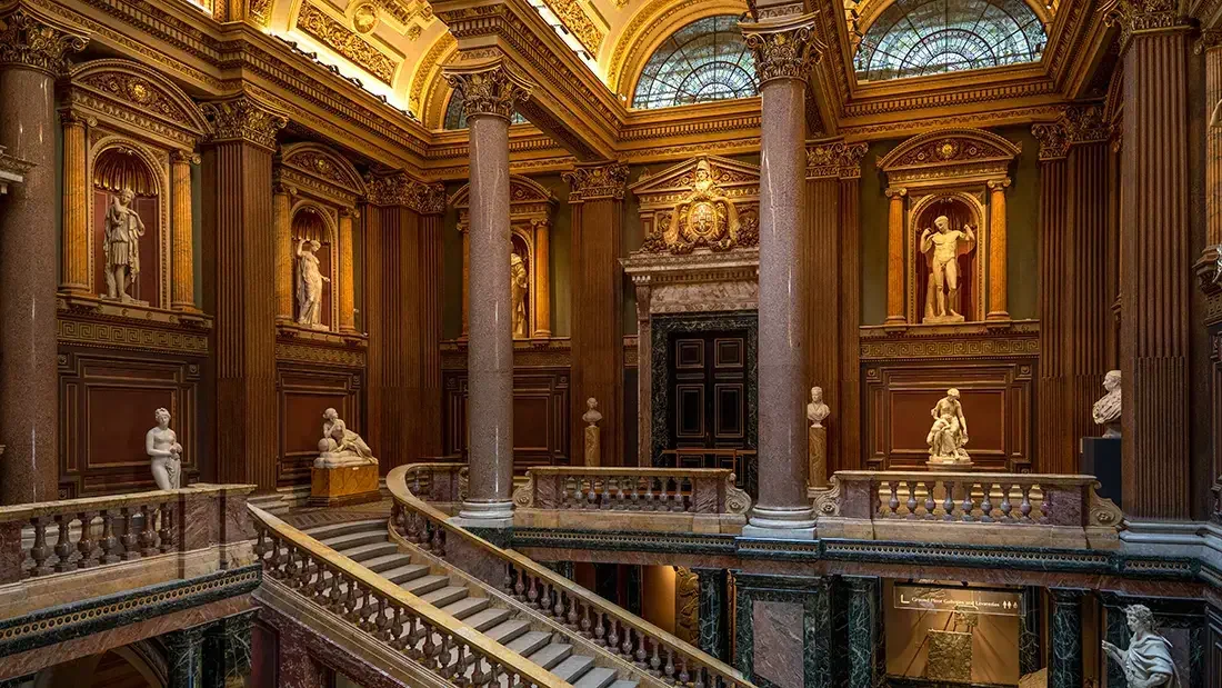 Interior of a grand classical building with a marble staircase, ornate columns, and a domed glass ceiling. The walls are decorated with statues in niches.