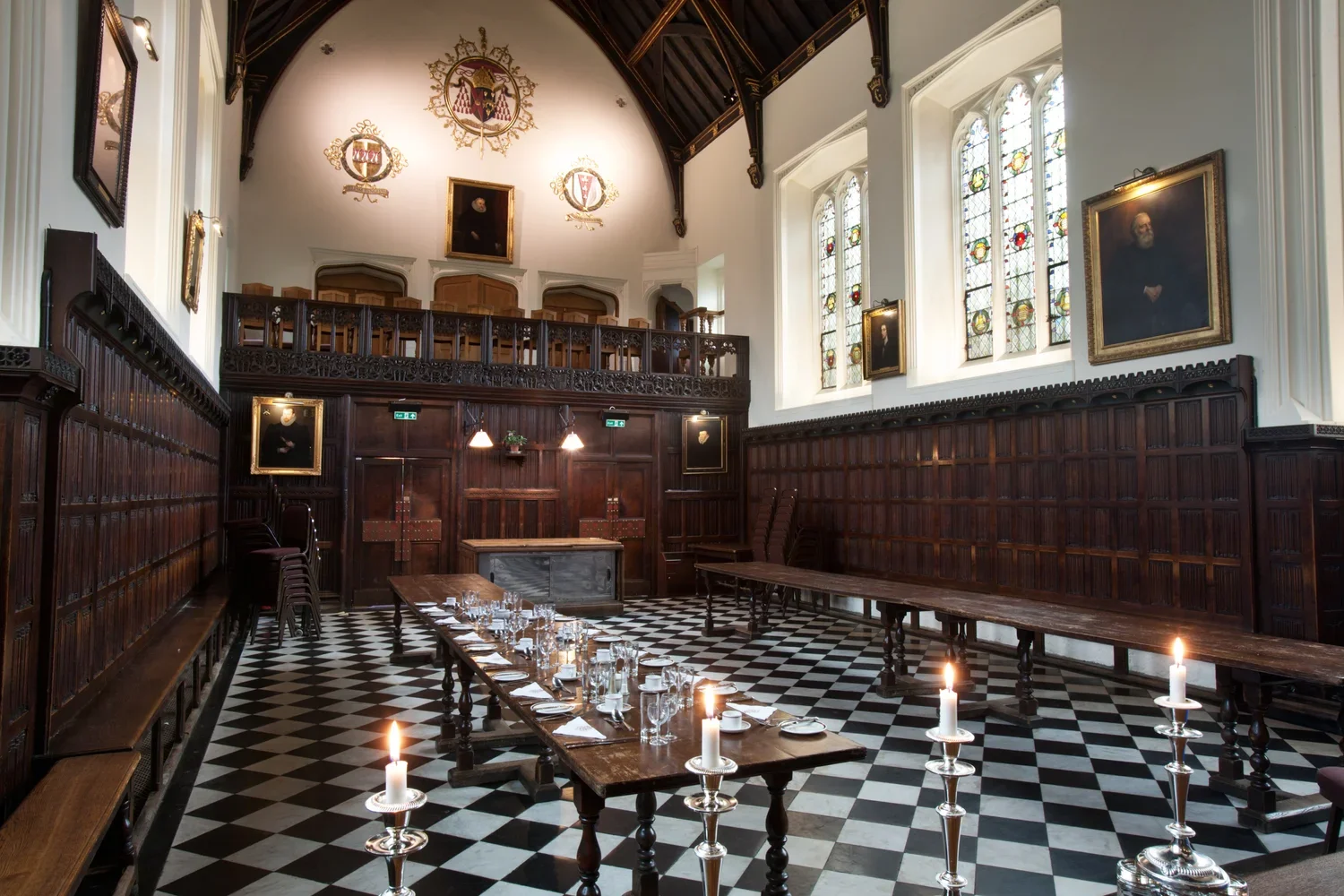 A historic, ornate room with high vaulted ceiling, stained glass windows, wooden paneling, and portraits on the walls, set up with long tables and lit candles, likely a dining hall or banquet room.