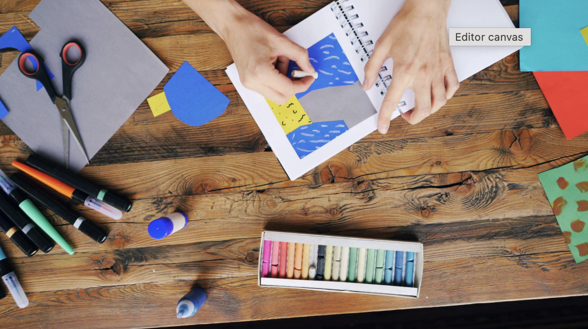 Hands creating a colorful collage with paper and markers on a wooden table, surrounded by art supplies including scissors, glue sticks, markers, chalk pastels, and colored paper.