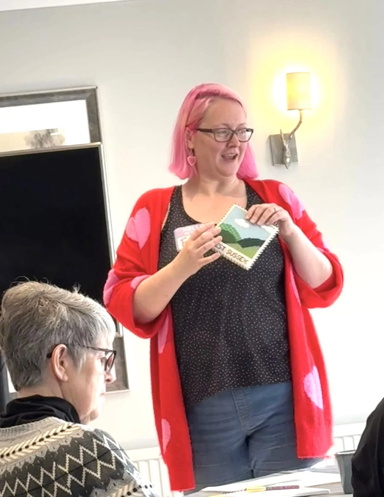 A woman with pink hair, glasses, wearing a red cardigan and earrings shaped like hearts, standing and speaking to a group, holding some papers or cards. Several people are seated, listening, with a plain white wall, a wall sconce, and a TV in the background.