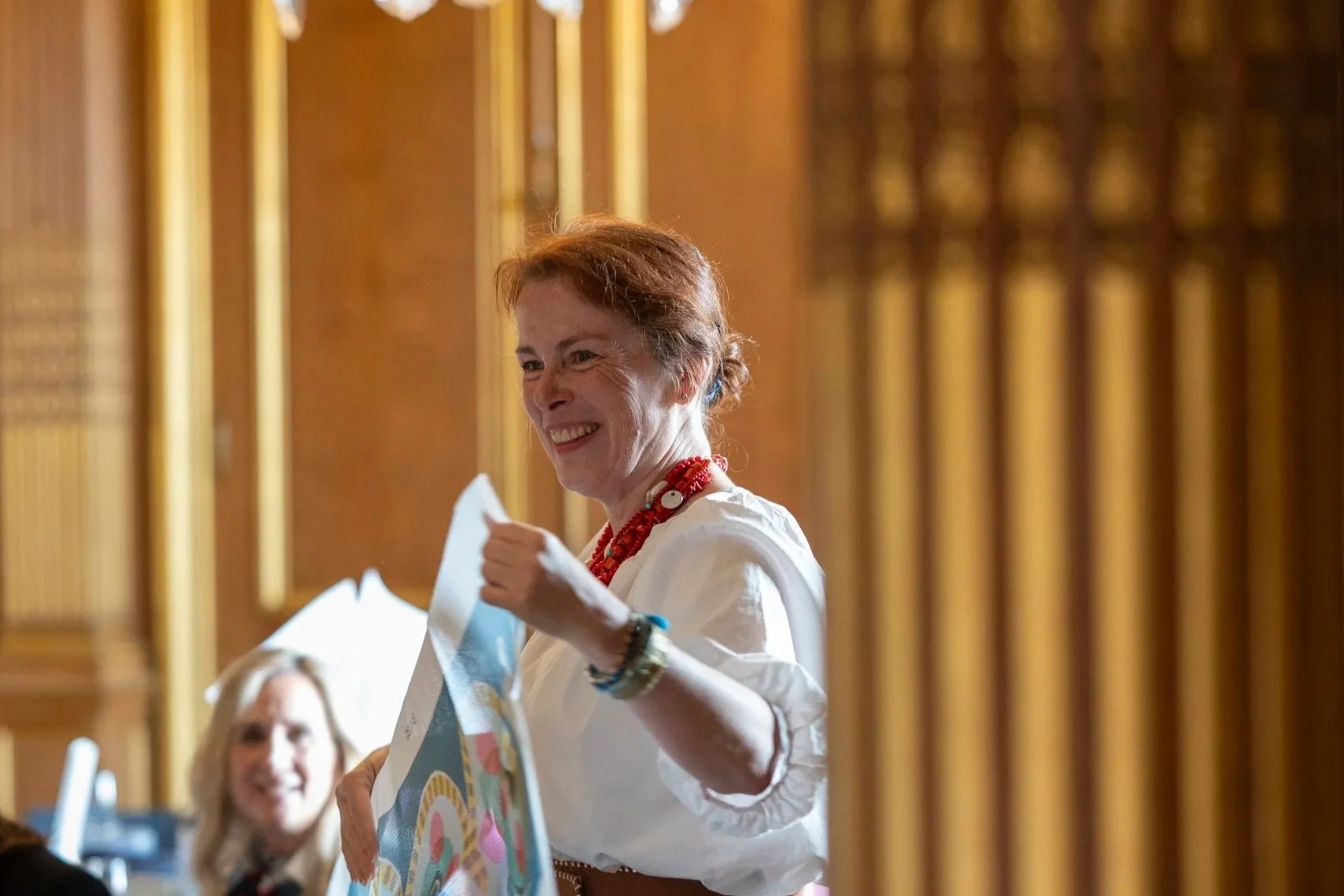 A woman with short red hair, wearing a white blouse and red necklace, smiling and holding a colorful object. There are wooden vertical bars in the foreground, and a blurred person is in the background.