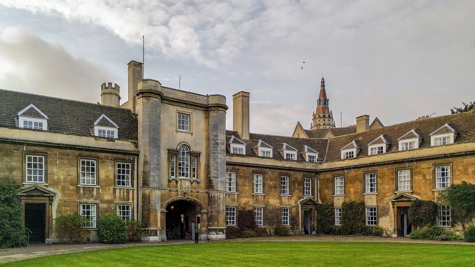Historic stone building with multiple windows and a central archway, surrounded by a well-kept lawn and shrubbery, with a church steeple in the background under cloudy sky.