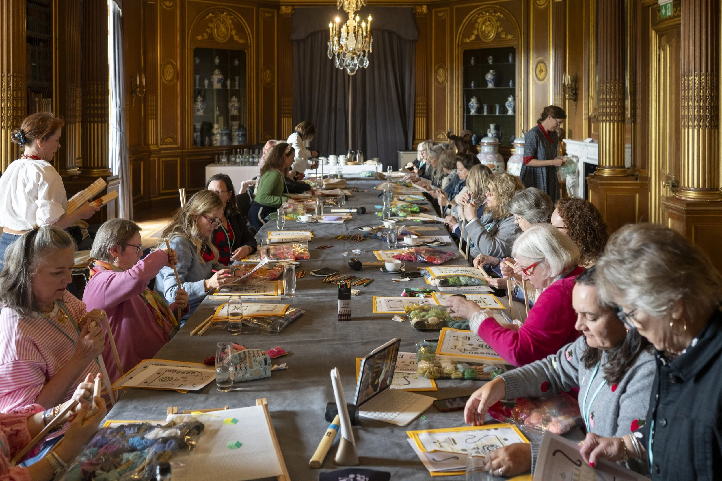 Women sitting at a long table in an ornate, wood-paneled room, engaging in a crafting activity with yarn, needles, and printed instructions, with some women standing and assisting others.