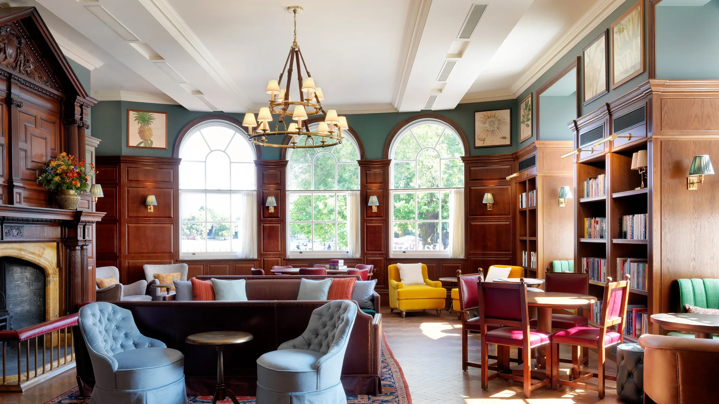 A spacious, well-lit library with large arched windows, wooden paneling, and a variety of colorful chairs and sofas arranged around. There is a chandelier hanging from the ceiling, framed botanical artwork on the walls, and bookshelves filled with books.