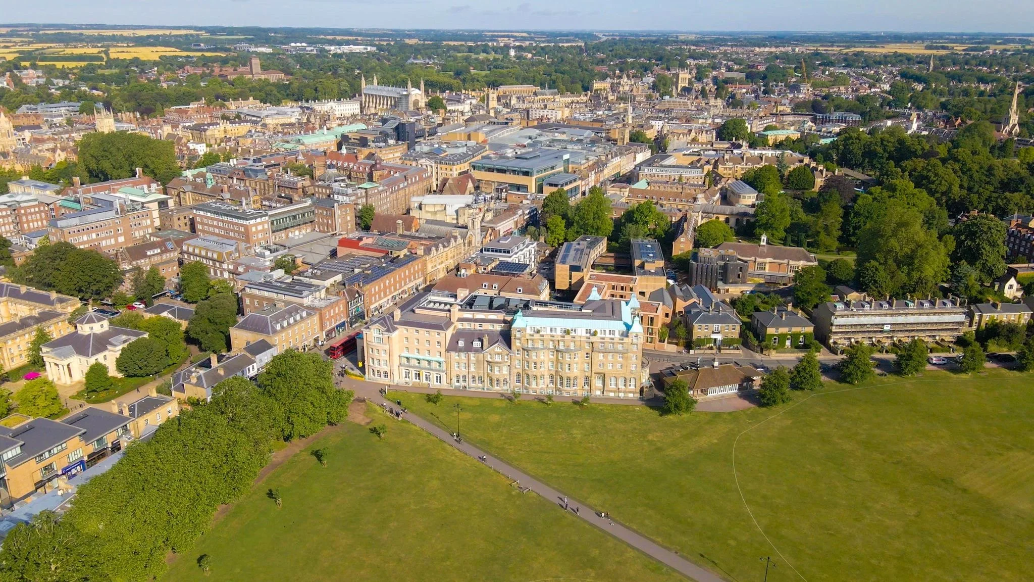 Aerial view of a cityscape with a mix of historic and modern buildings, lush green trees, and sports fields in the foreground.