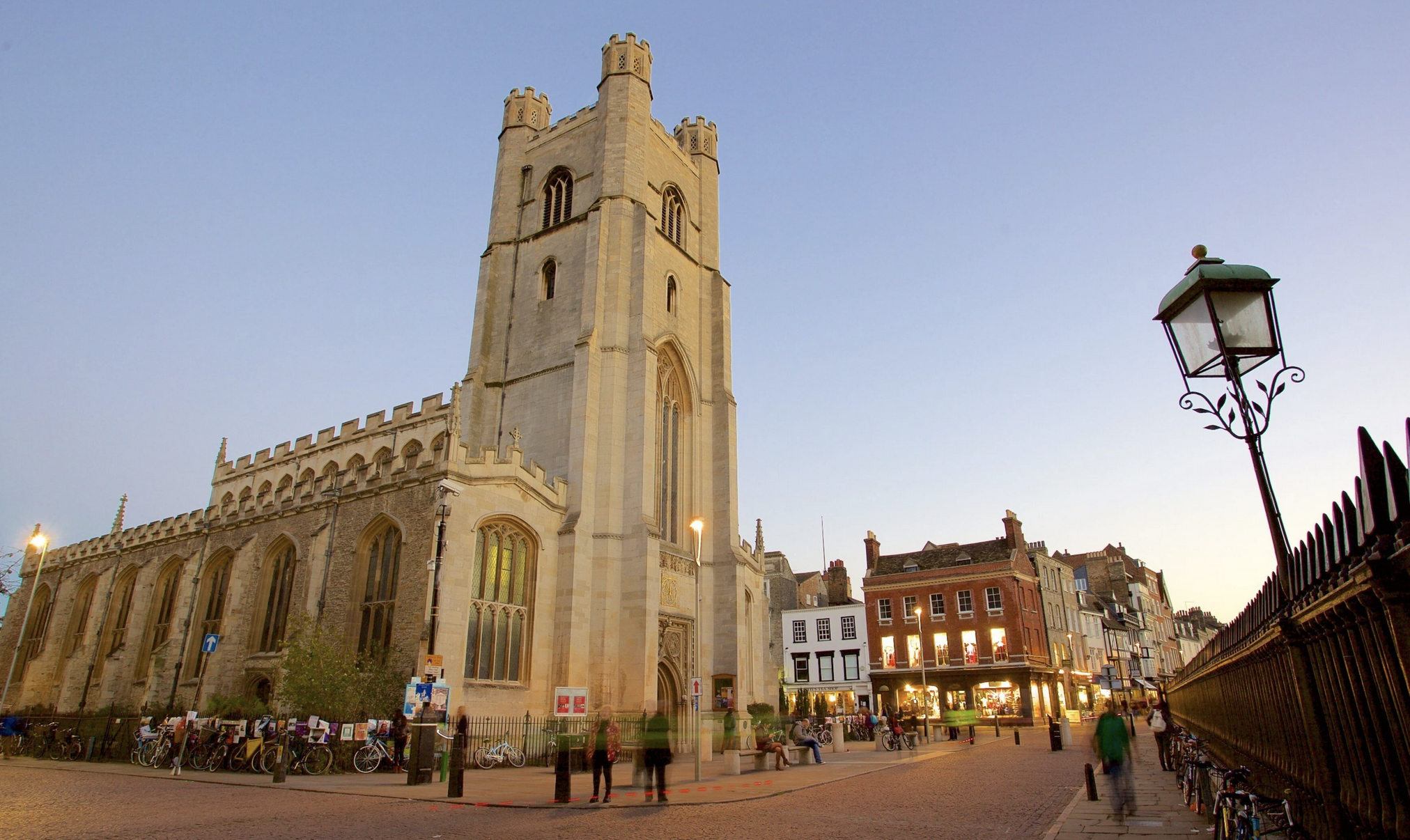 A historic church with a tall stone tower and arched windows, illuminated by streetlights, in a city square with bicycles parked nearby, surrounding buildings, and a clear evening sky.