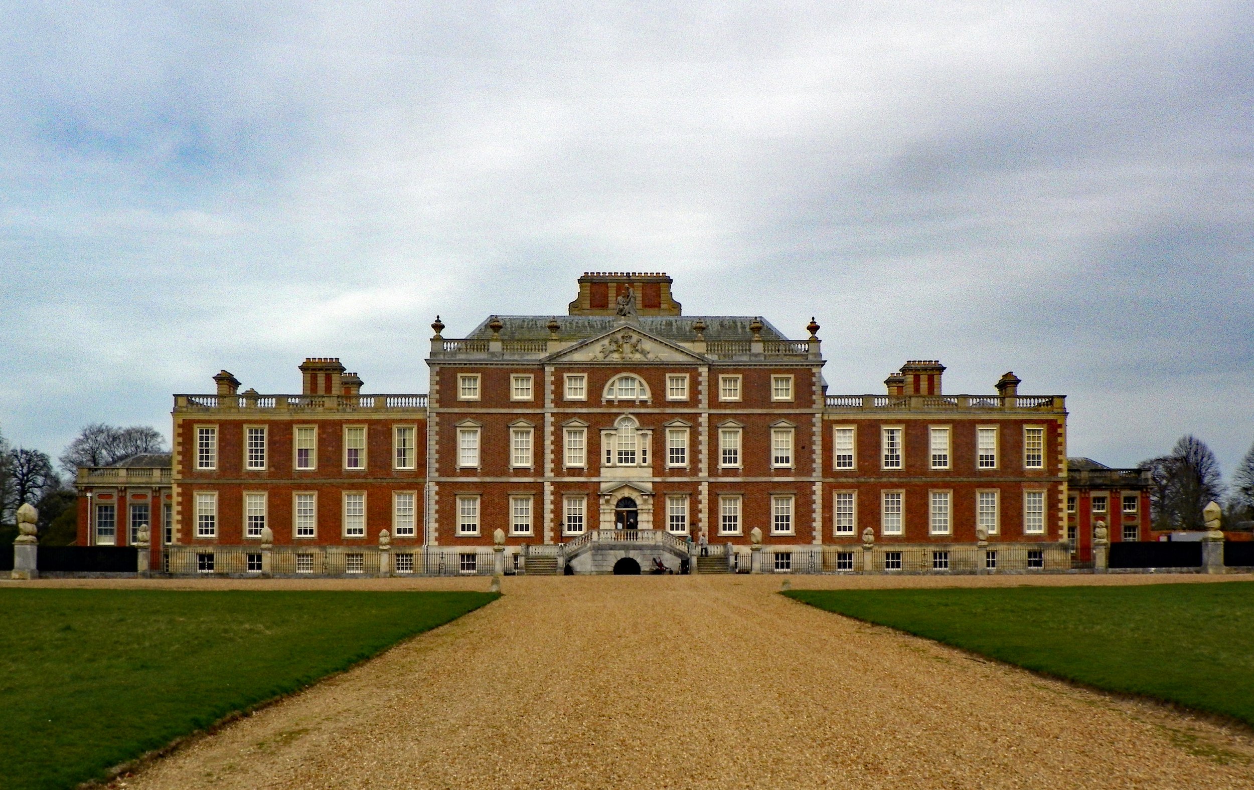 A grand red-brick mansion with multiple windows and decorative architectural details, surrounded by a green lawn and a gravel pathway leading to the entrance under a cloudy sky.