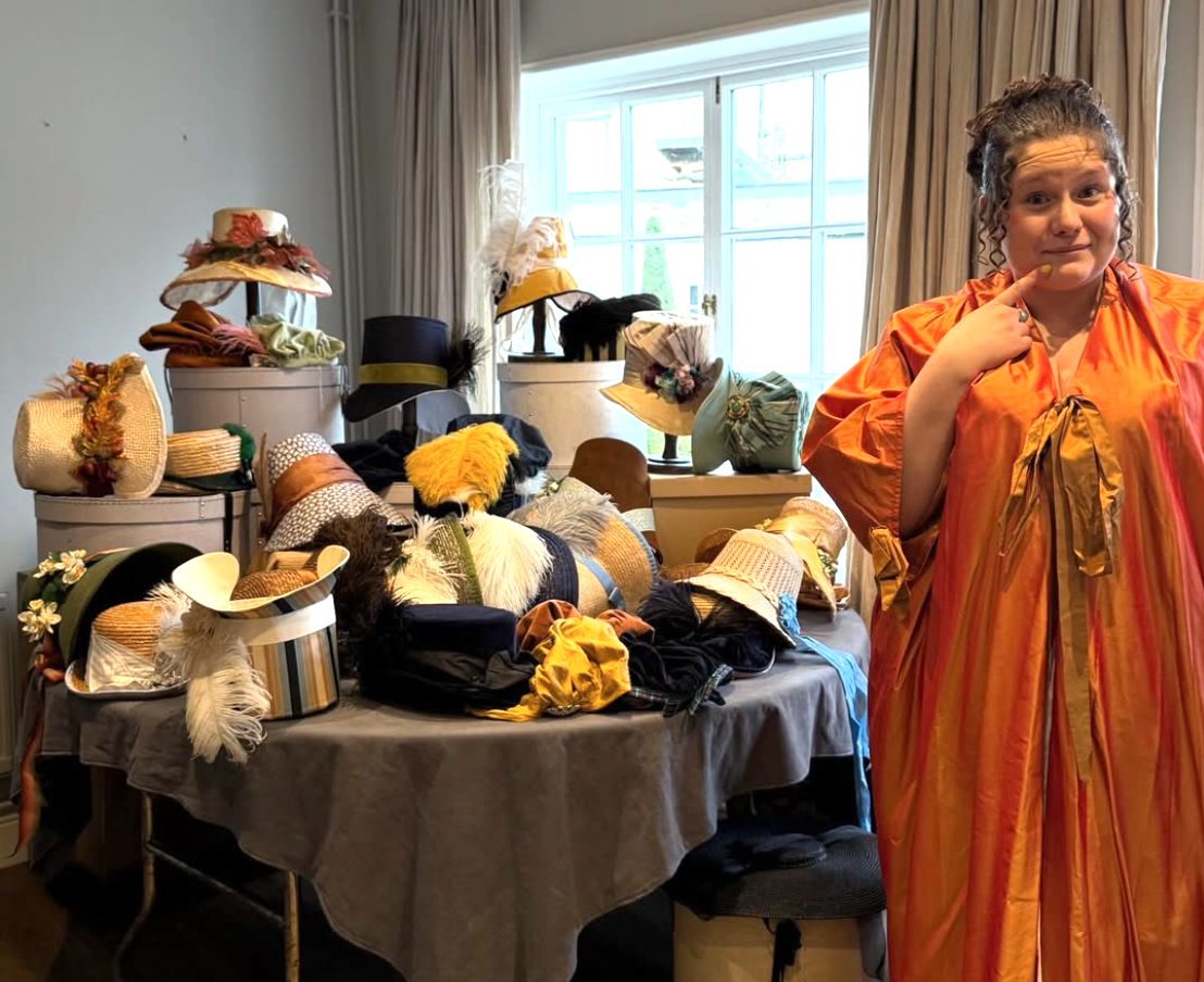 A woman standing next to a table filled with various hats, some decorated with feathers and flowers, in a room with large windows and beige curtains.