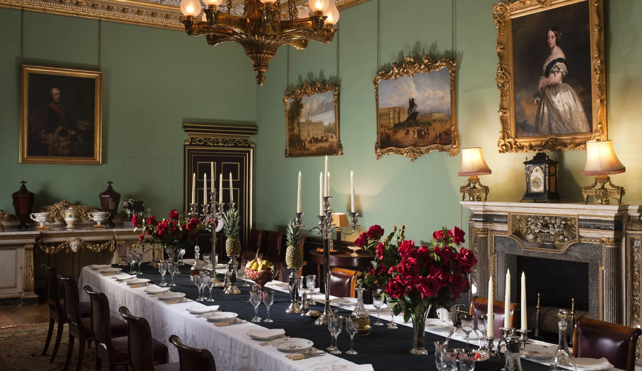 A luxurious dining room with a long table set with fine china, crystal glasses, and silverware, surrounded by dark wooden chairs. The room features elegant green walls, ornate gold-framed paintings, a chandelier, and a fireplace decorated with gold accents. Fresh flowers and candles decorate the table.