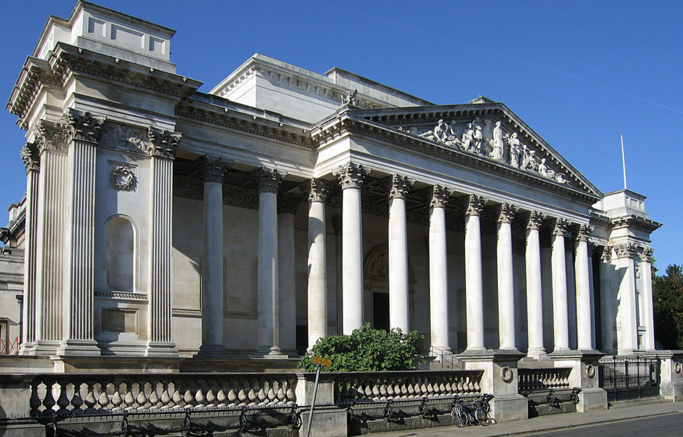White classical building with tall columns, decorative friezes, and a triangular pediment with sculpted figures, set against a blue sky.