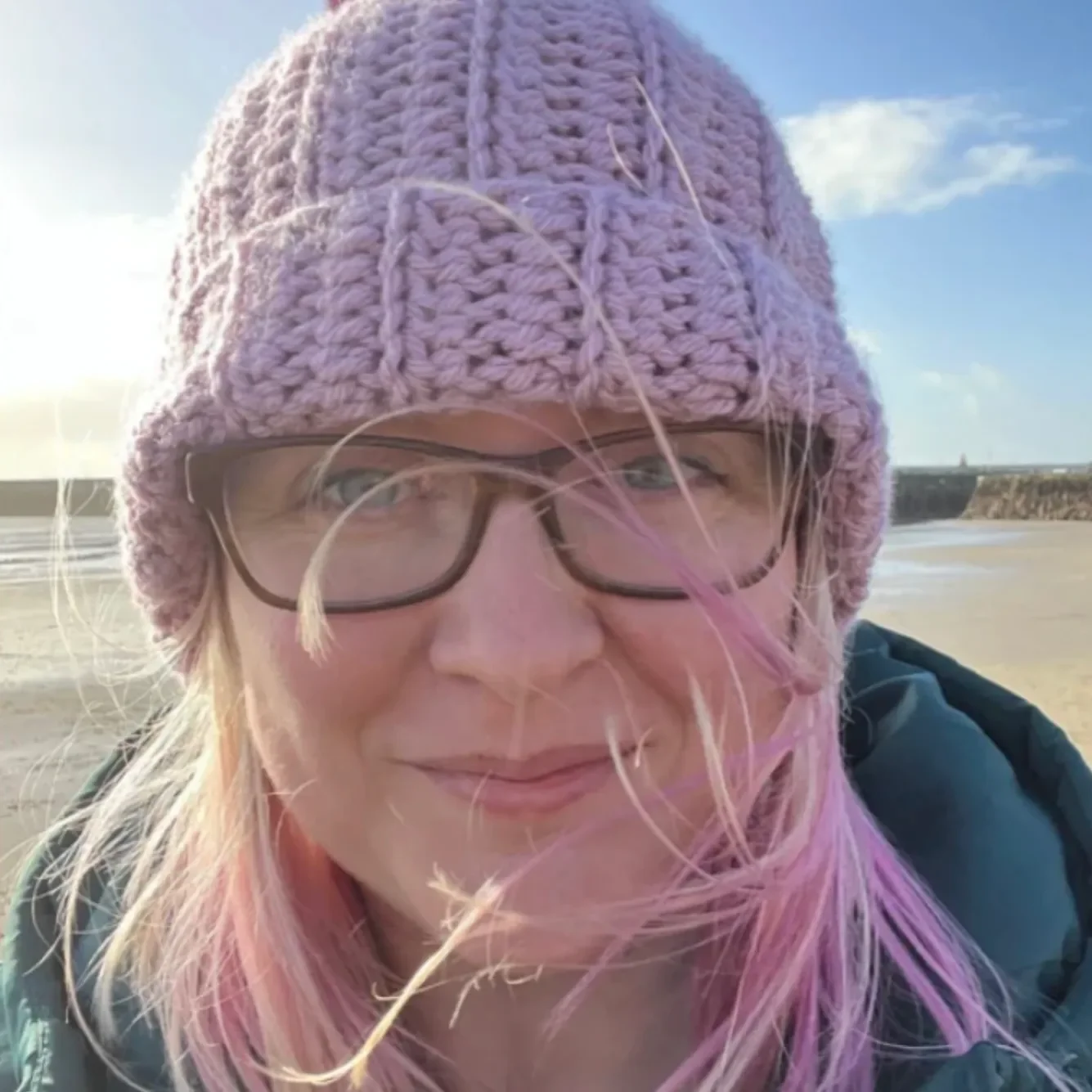 A woman with pink hair wearing glasses and a knitted pink hat, outdoors on a beach with a clear sky and distant cliffs in the background.