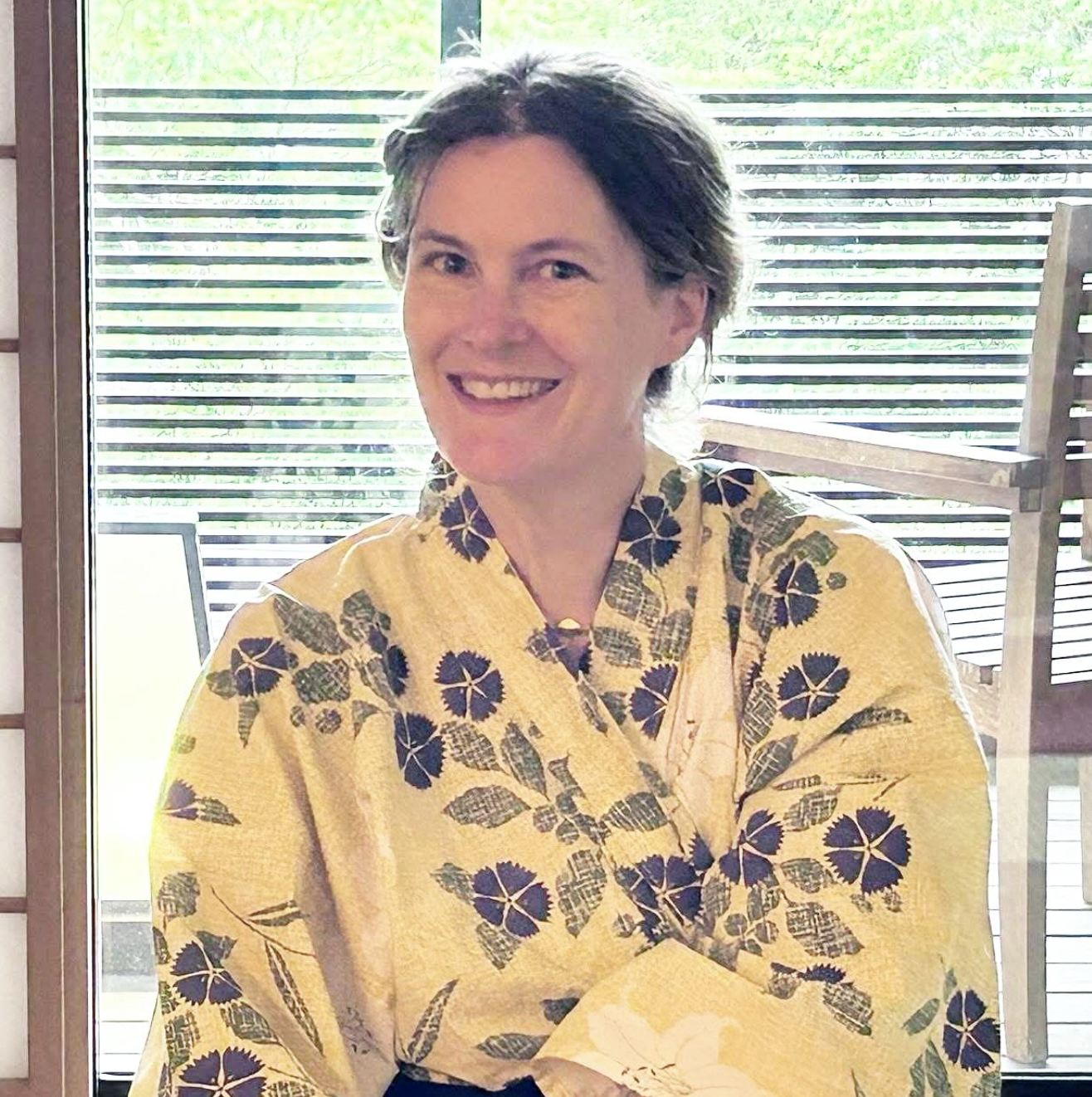 A woman smiling while sitting indoors, wearing a yellow kimono with purple and green floral patterns, with a background of green trees visible through the window.