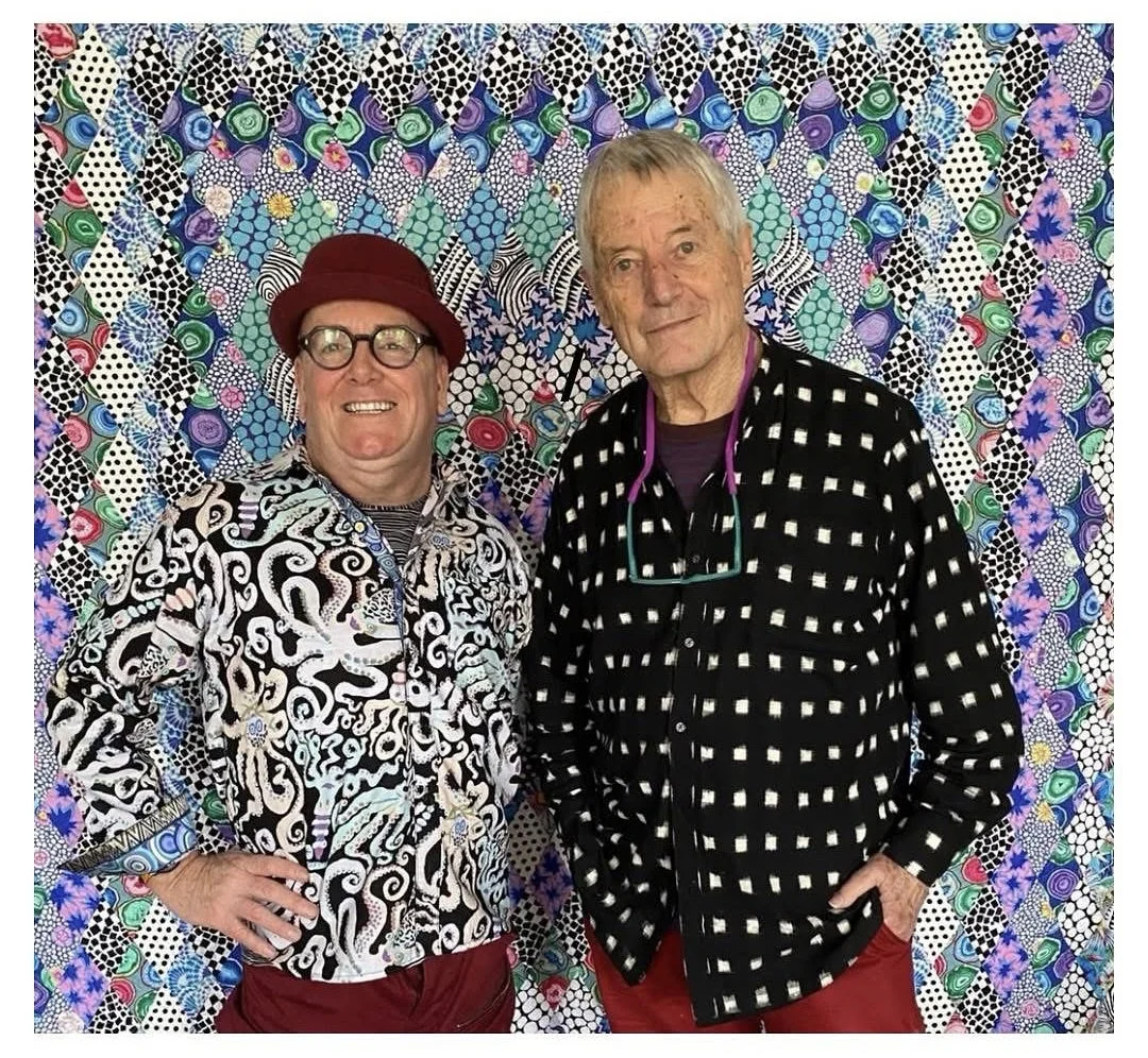 Two men standing in front of a colorful, patterned quilt display. The man on the left wears glasses, a red hat, a black and white patterned shirt, and red pants. The man on the right has gray hair, wears sunglasses hanging from his shirt, a black and white checkered shirt, and red pants.