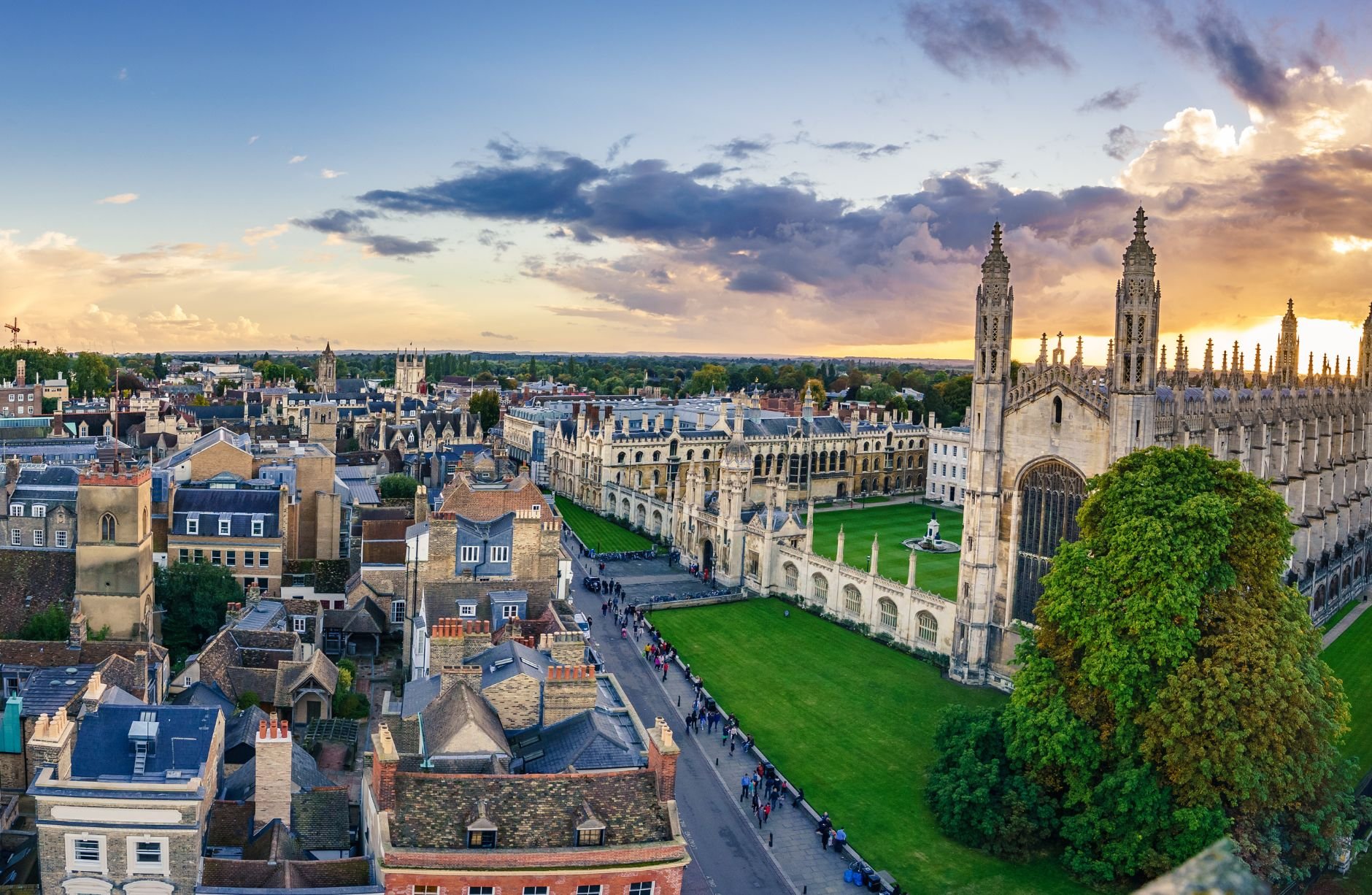 A panoramic view of a historic European city with a large Gothic cathedral, green lawns, and a street filled with people during sunset.