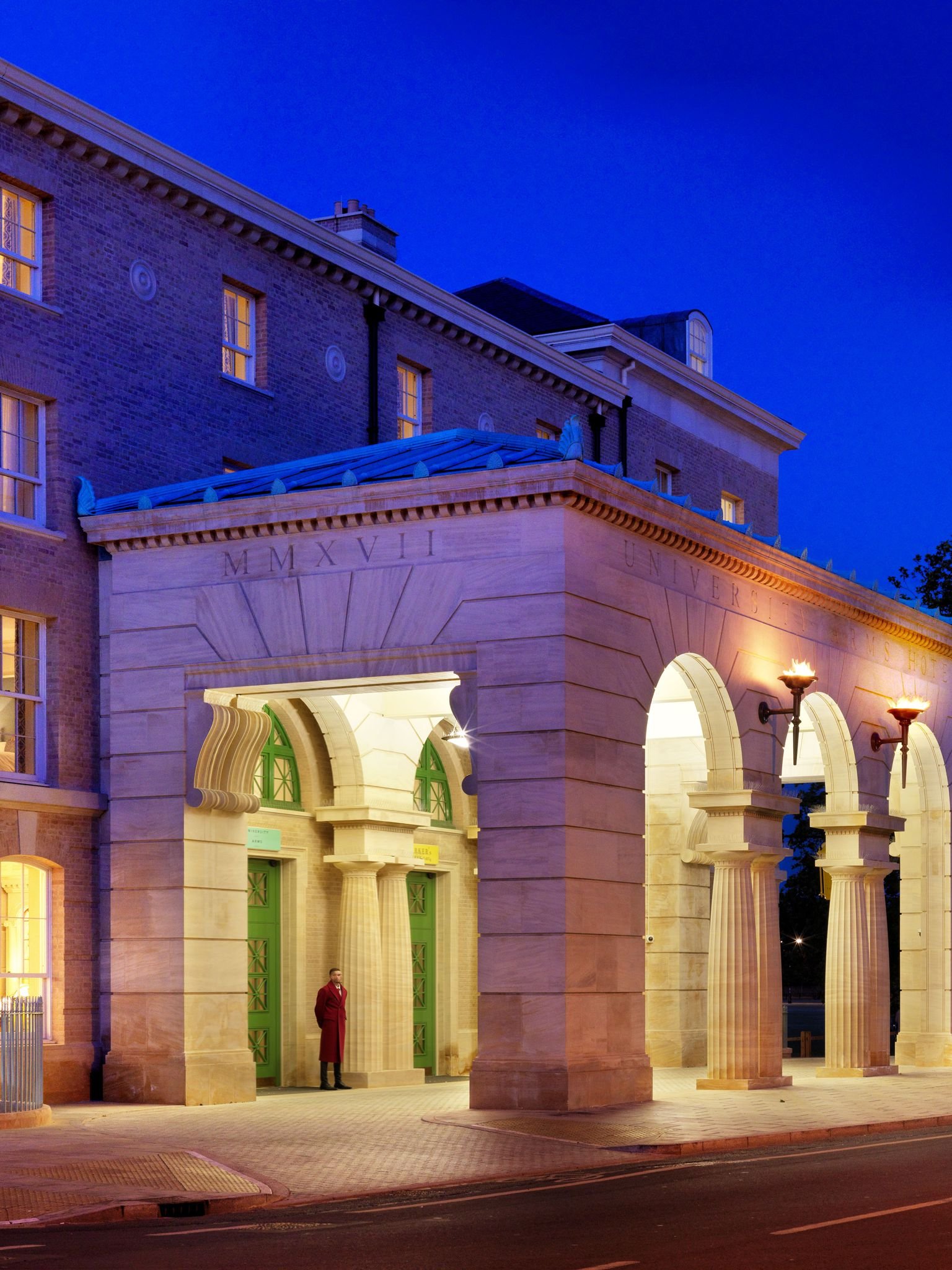 A building with classical architectural elements like columns and a large entrance, illuminated during the evening. The building bears the words 'UNIVERSITY' and Roman numerals 'MMXVII' above the entrance, with a man in a red coat standing near the door.