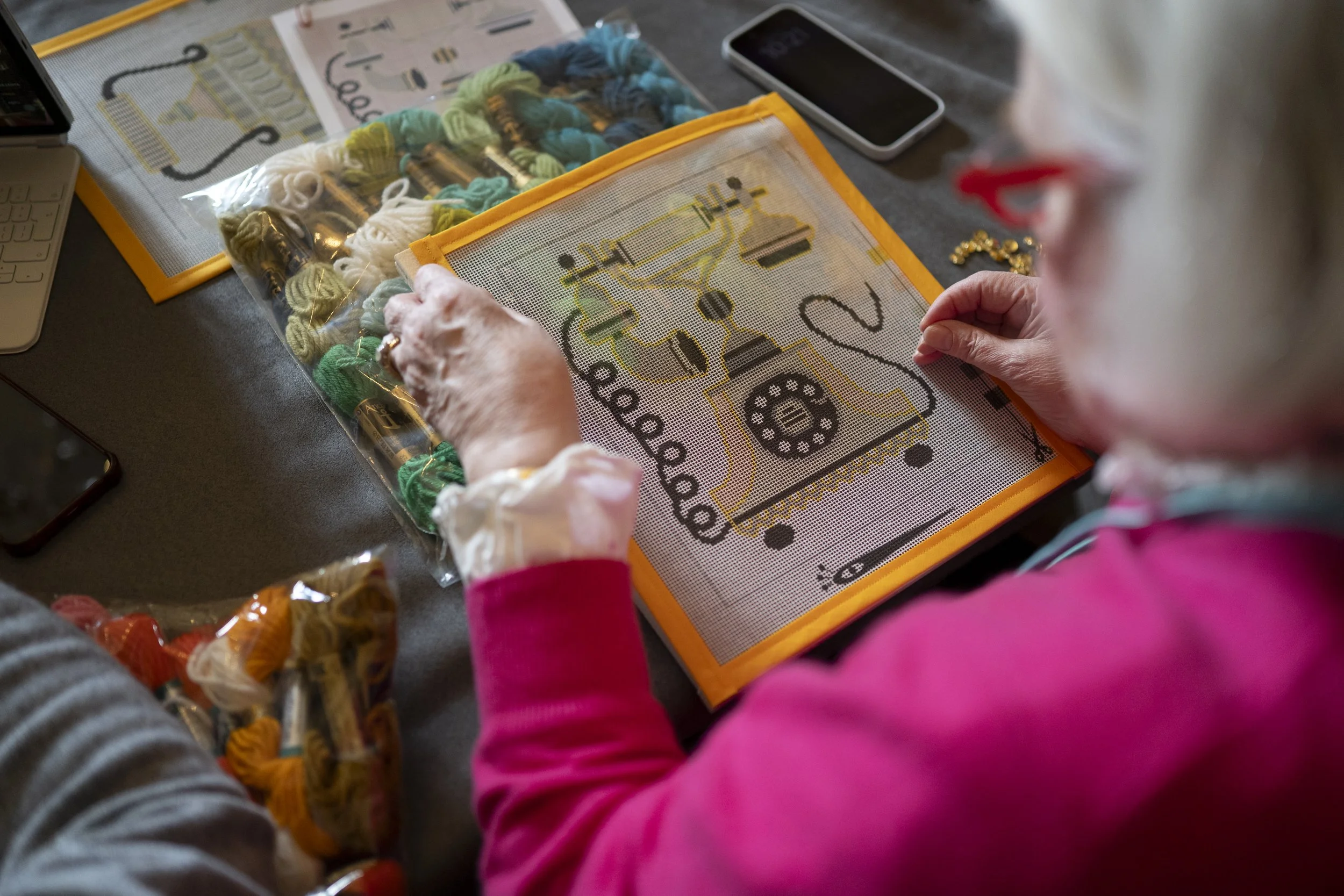Child with pink sleeves working on a needlepoint craft project on a table, with skeins of yarn, a smartphone, and a partially completed cross-stitch picture of a telephone.