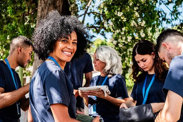 Group of healthcare workers in scrubs outdoors with trees, some looking at tablets and smiling.