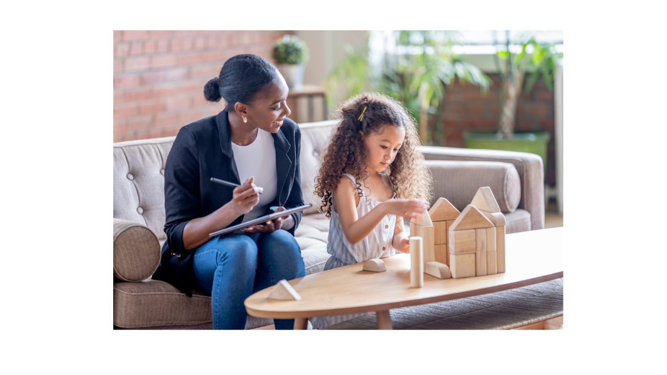 A woman and a young girl sitting on a sofa in a bright room, engaging in a play activity with wooden building blocks on a table.