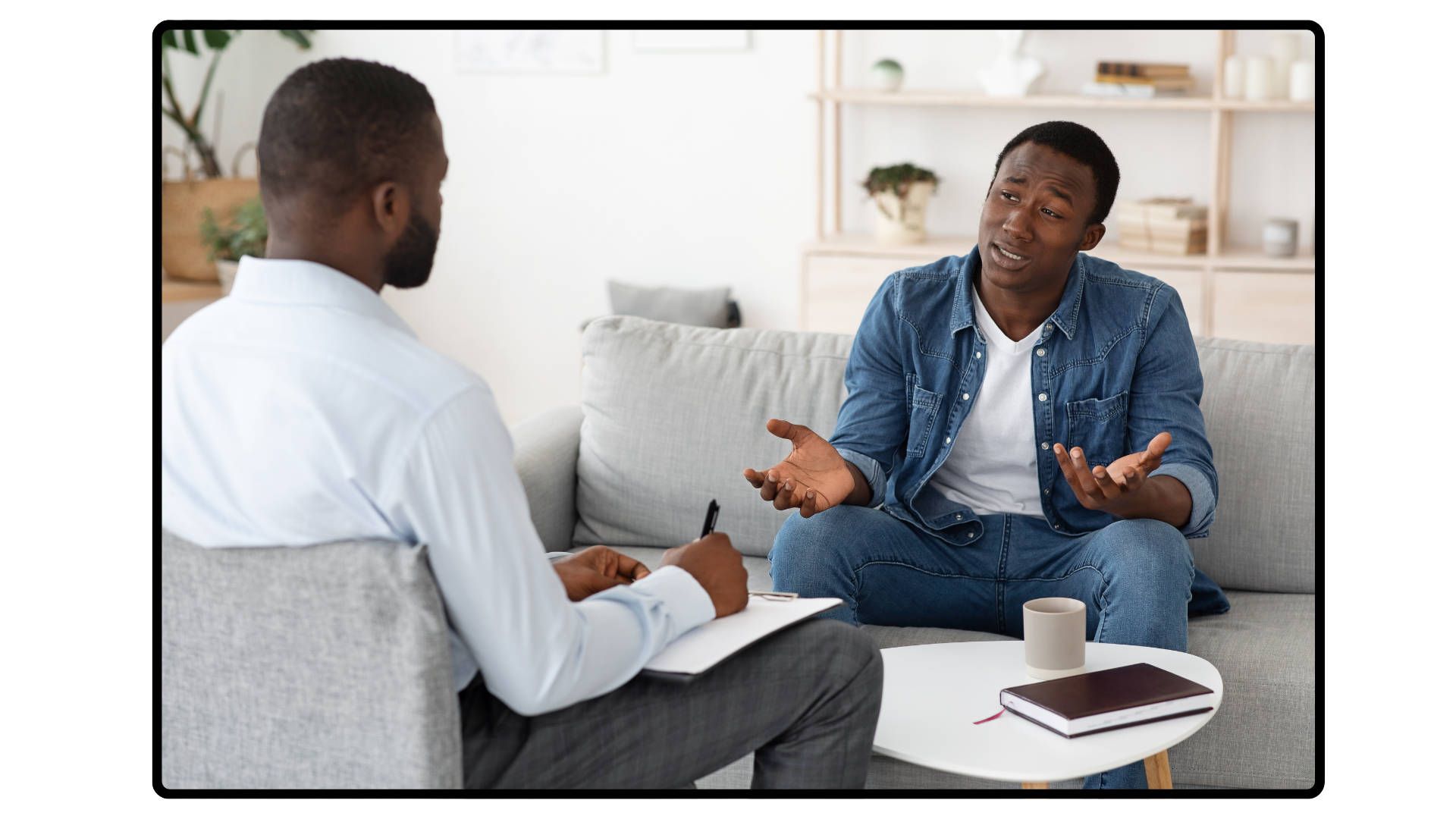 Two men sitting on a sofa having a conversation, one man is gesturing with his hands. There is a coffee table in front of them with a notebook, a pen, a cup, and a tablet. The setting appears to be a living room with shelves and decorative items in the background.