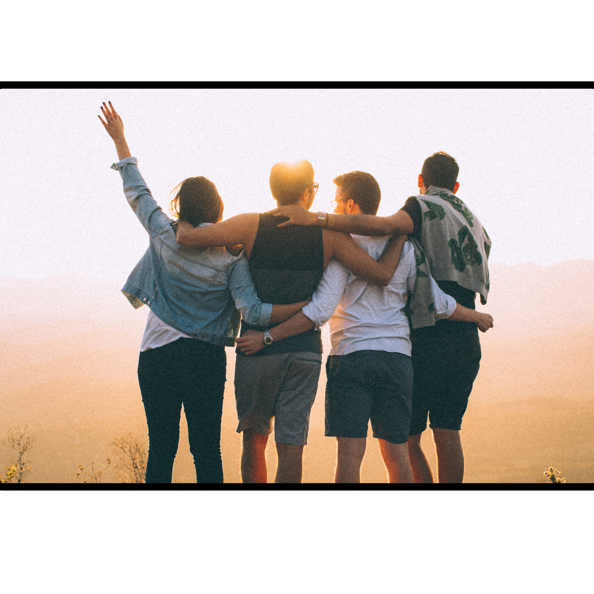 A group of five friends hugging and sitting together outdoors during sunset, with one person raising their hand.