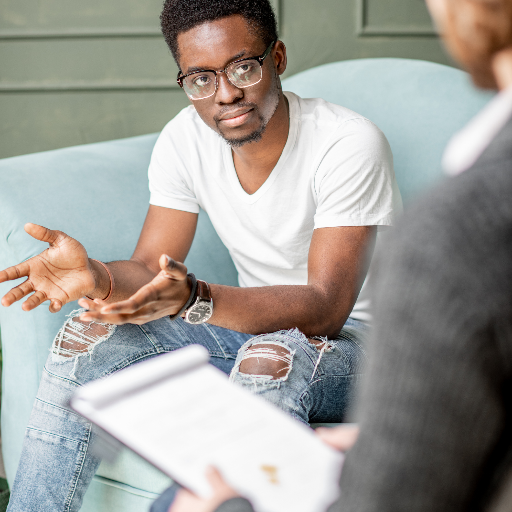 A young man with glasses and a white T-shirt is sitting on a pastel green couch, looking at a woman who is sitting across from him, partially visible in the foreground. They appear to be having a conversation.
