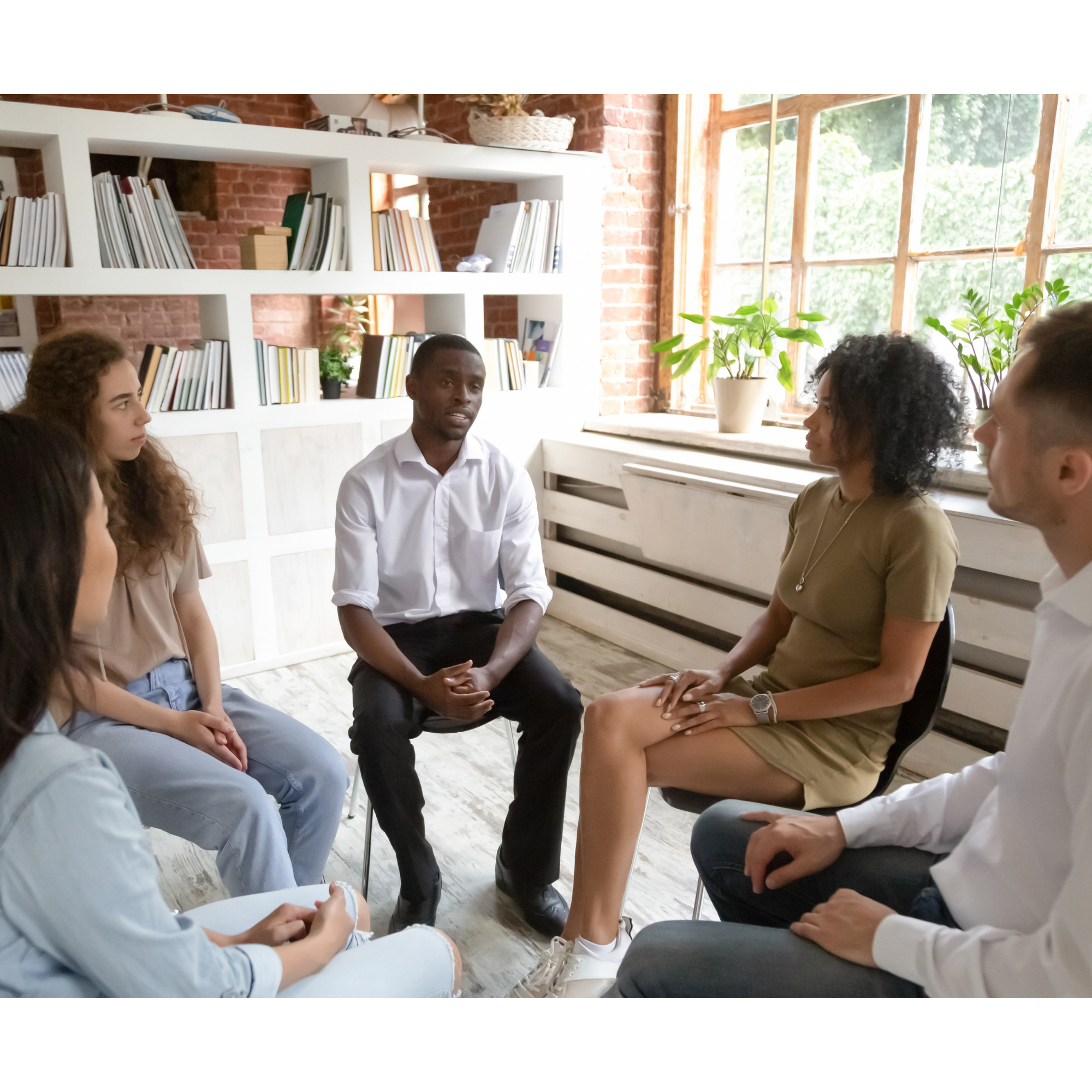 Group of diverse young adults sitting in a circle having a discussion in a bright room with large windows and a bookshelf in the background.