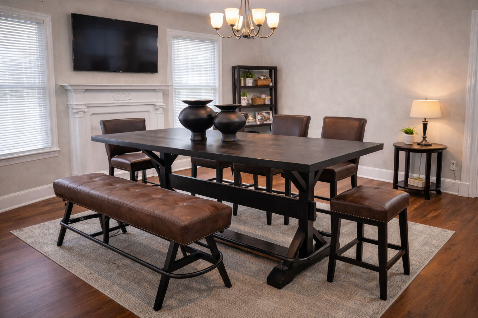 A dining room with a dark wooden table, brown leather chairs, a brown leather bench, and a beige rug. There are black vases on the table, a flat-screen TV above a white fireplace mantel, a small bookshelf with decorative items, a side table with a lamp and small plant, and windows with white blinds.