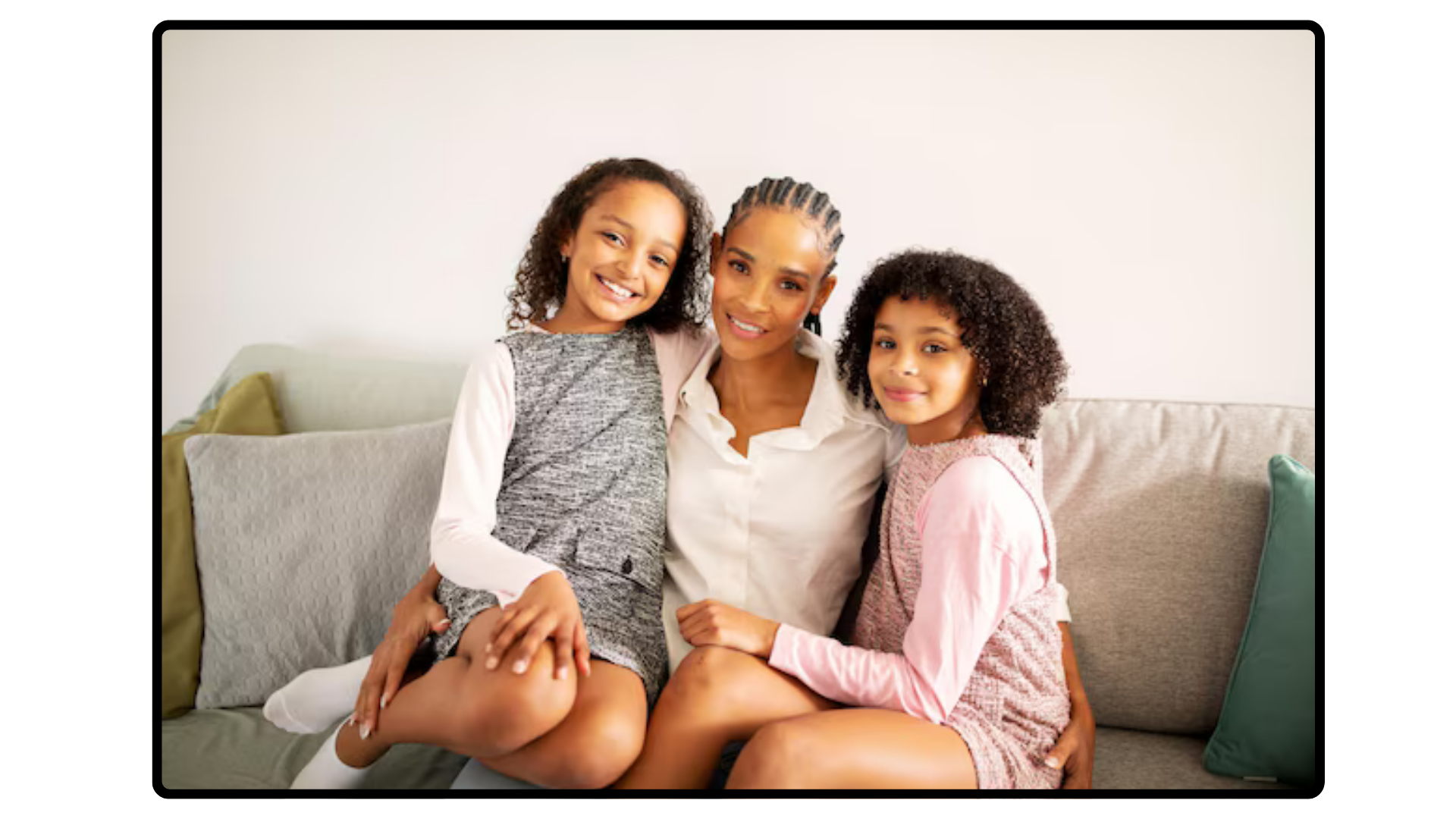 A woman sitting on a sofa with two young girls, all smiling and looking at the camera. The woman has braided hair and is wearing a white shirt. The girl on the left has curly hair and is wearing a gray and white dress. The girl on the right has curly hair and is wearing a pink and white outfit. There are pillows on the sofa and a plain white wall in the background.