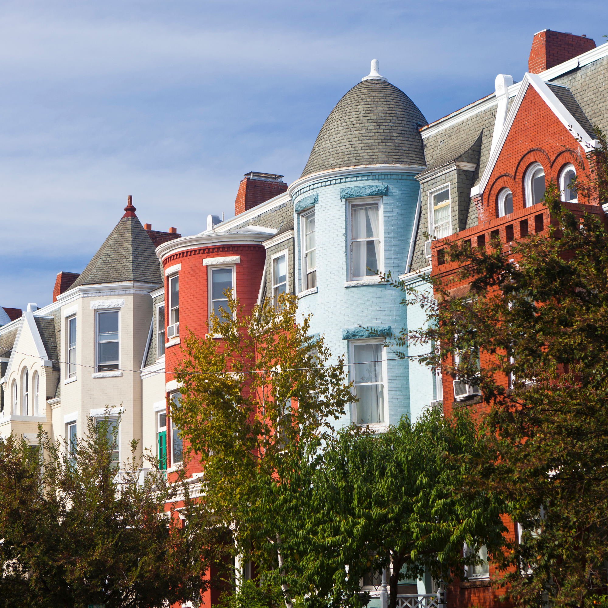 Colorful Victorian-style houses with unique turrets and gabled roofs, partially obscured by trees, under a blue sky.