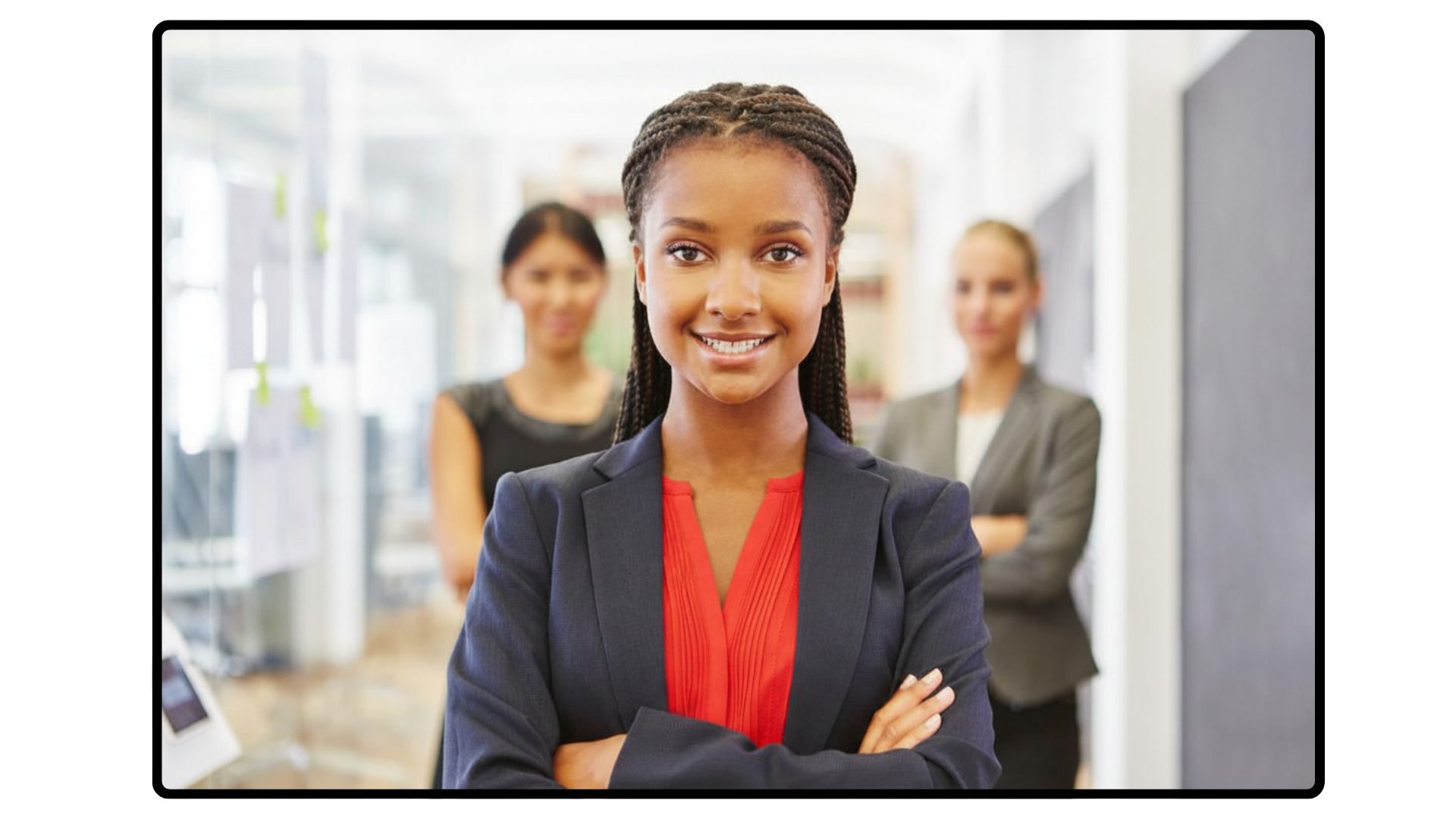A woman with braided hair, wearing a navy blazer and red blouse, smiling with arms crossed in an office or conference setting. Two women are in the background, slightly blurred.