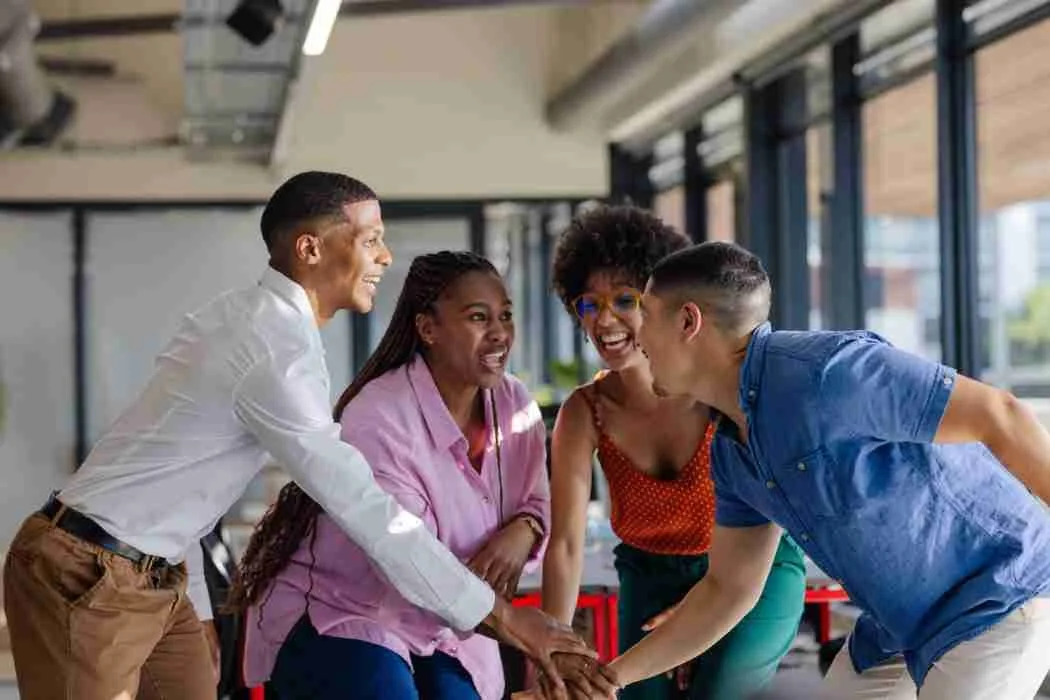 Four diverse coworkers smiling and high-fiving in an office with large windows.