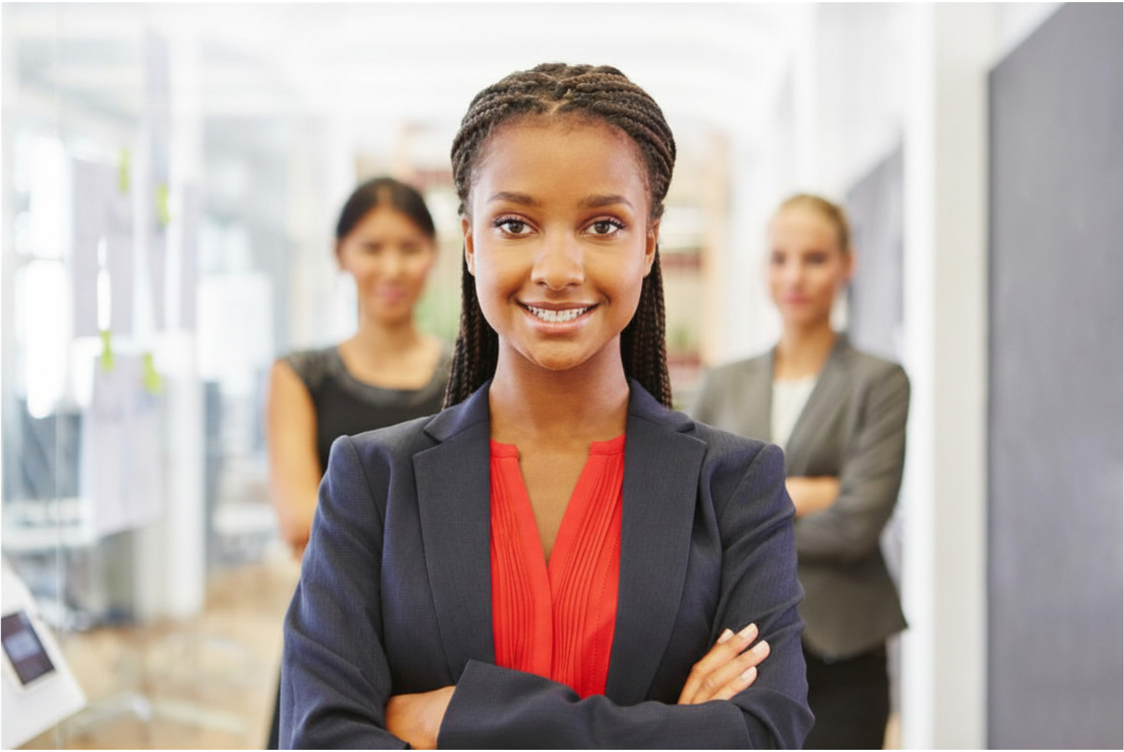 A confident African American woman in business attire standing with arms crossed, smiling, with two women blurred in the background in a professional office setting.