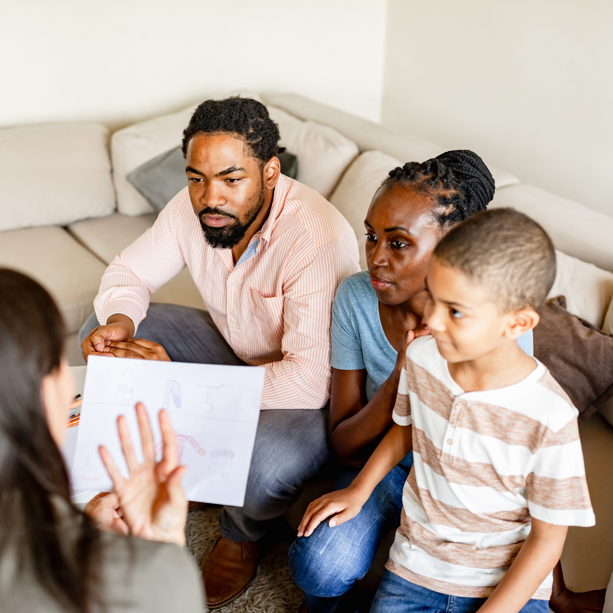 Group of children and adults sitting on a beige couch, participating in a discussion or activity, with a person in the foreground holding a sheet of paper.