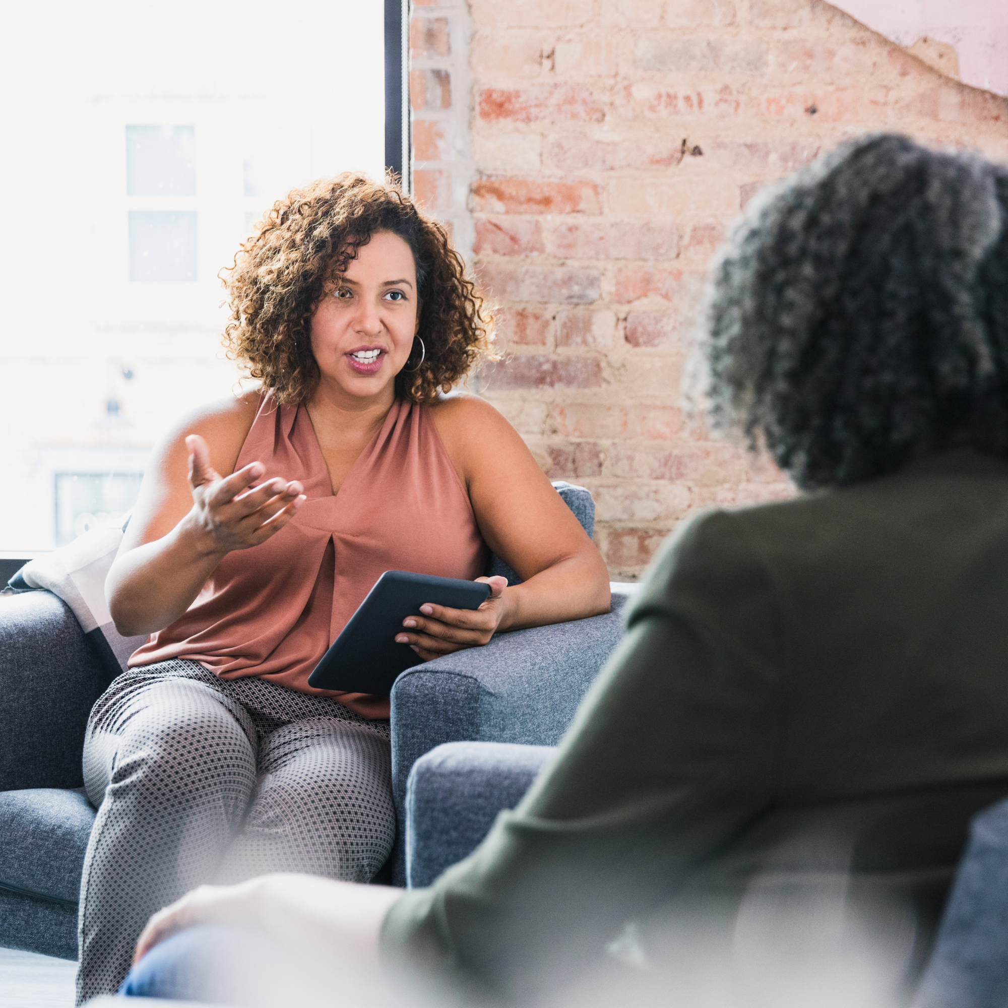 A woman with curly hair and hoop earrings talking during a conversation, holding a smartphone, sitting on a couch in a room with exposed brick wall.
