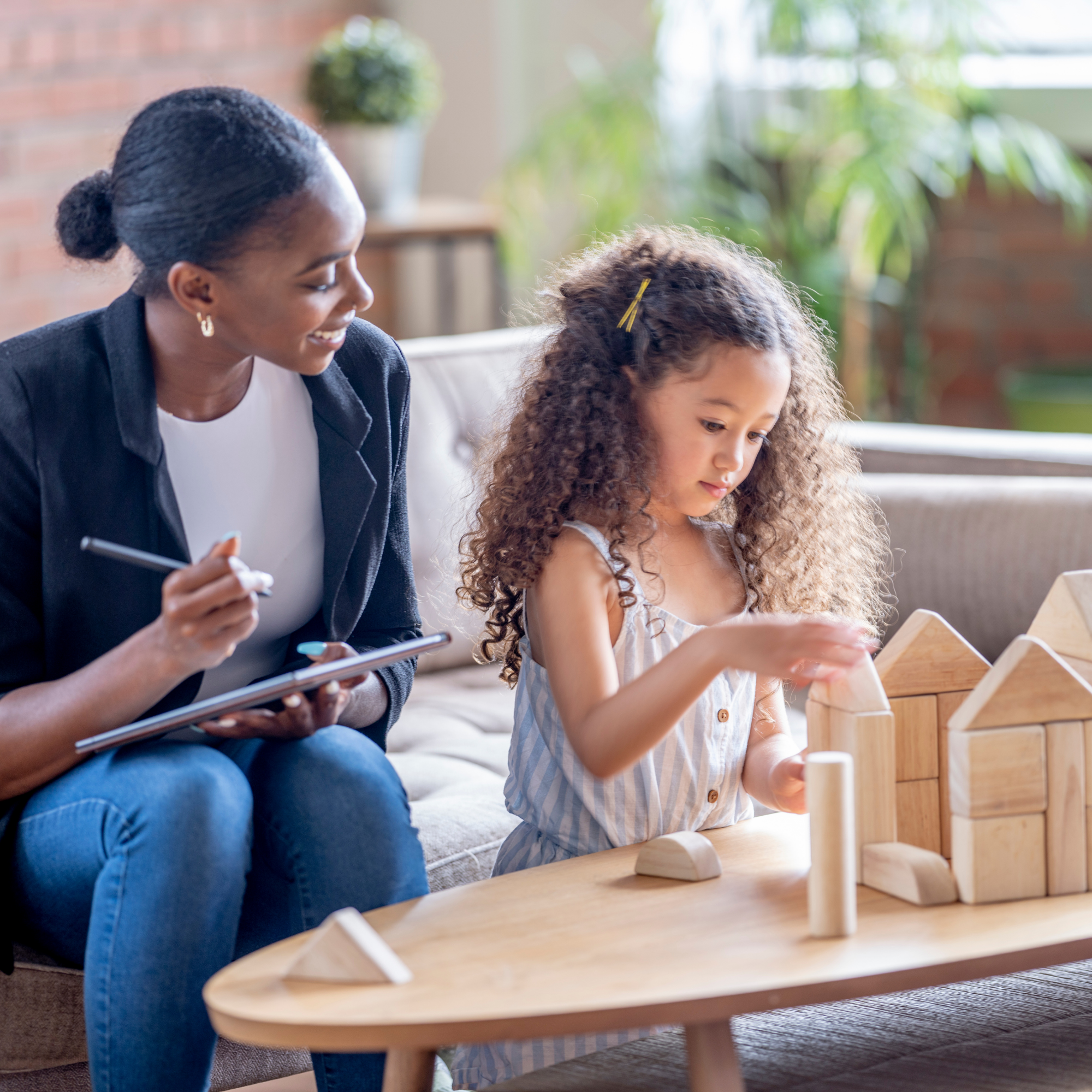 A woman with dark hair and earrings smiling while sitting next to a young girl with curly hair, engaged in playing with wooden toys at a table in a well-lit room with green plants in the background.